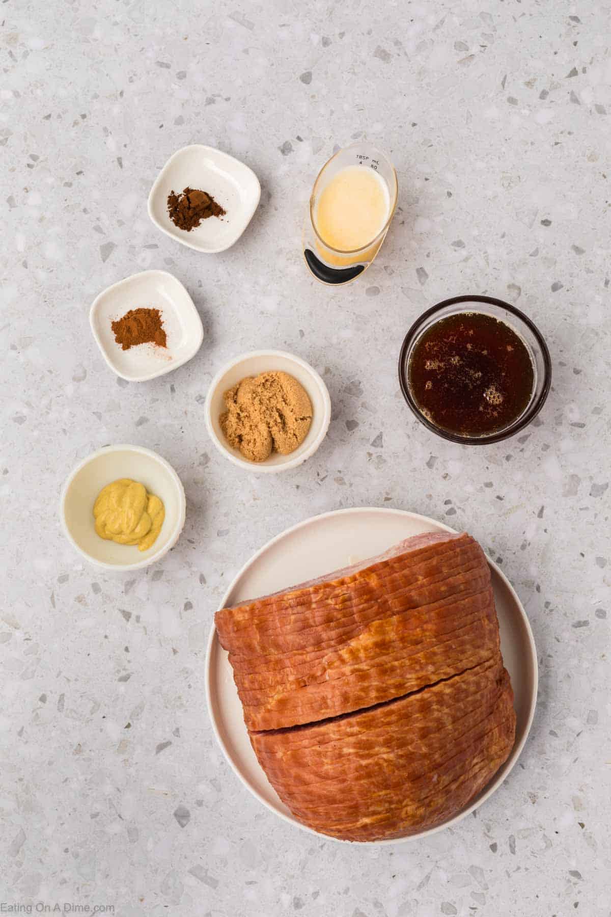 A sliced ham is artfully arranged on a white plate, surrounded by small bowls of brown sugar, mustard, ground spices, and a glass of dark liquid on the speckled countertop—perfect for an air fryer feast.