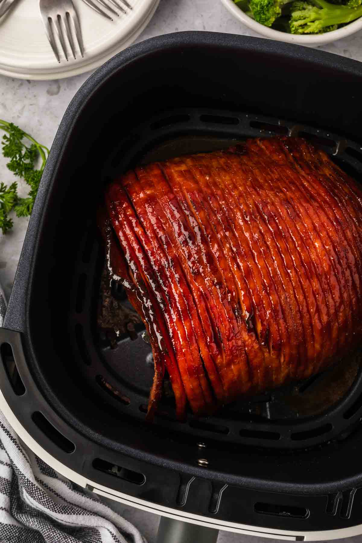 A delicious glazed ham sizzles to perfection in the air fryer basket, its surface caramelized and tempting. Forks and plates await in the background, accompanied by a fresh sprig of parsley on a light-colored surface.