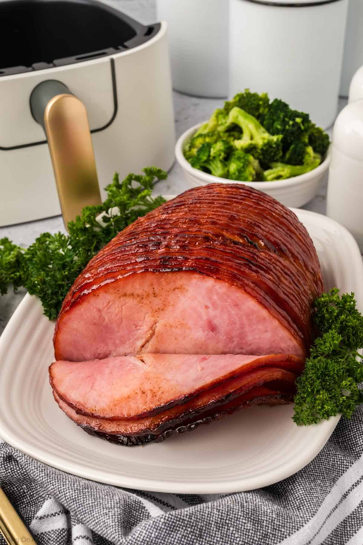 A succulent, air fryer-glazed ham rests on a white platter garnished with parsley. Broccoli pieces appear in a small white bowl in the background. A gold-handled utensil and a striped cloth complete the inviting tablescape beside the dish.