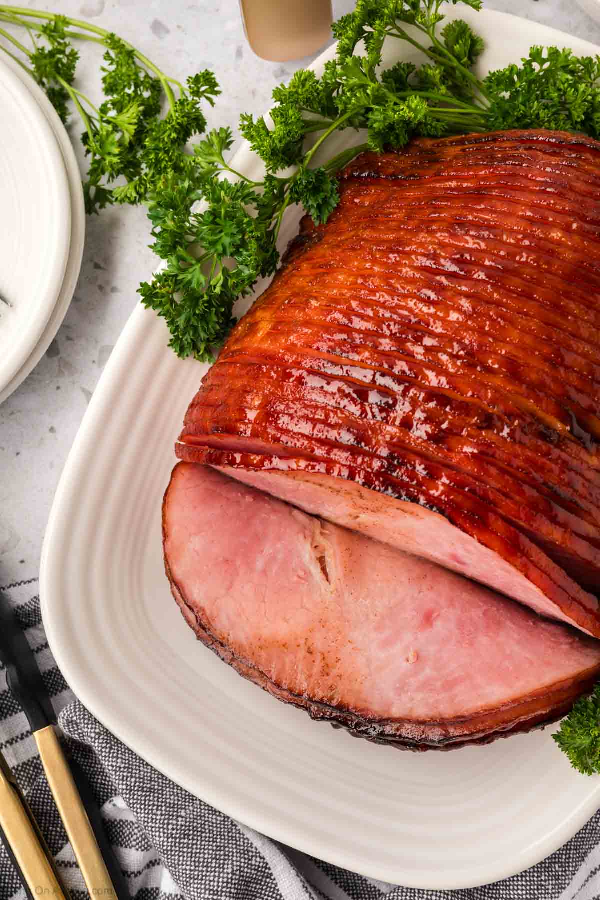 A glazed, spiral-sliced ham on a white platter, garnished with fresh parsley, shines with a caramelized appearance. Cooked to perfection in an air fryer, it sits enticingly on a light countertop beside a striped cloth and utensils.