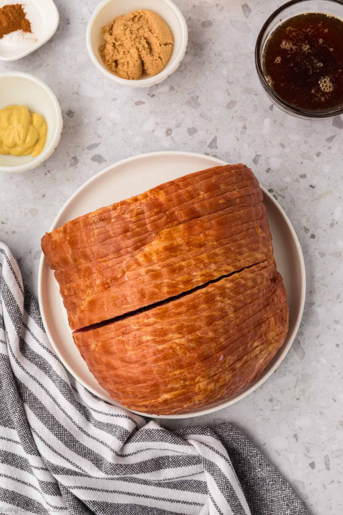 A glazed ham sits on a white plate, surrounded by small bowls of brown sugar, mustard, and spices on a speckled countertop. Perfect for an easy recipe, an air fryer can simplify preparing this dish. A striped cloth napkin is draped nearby.