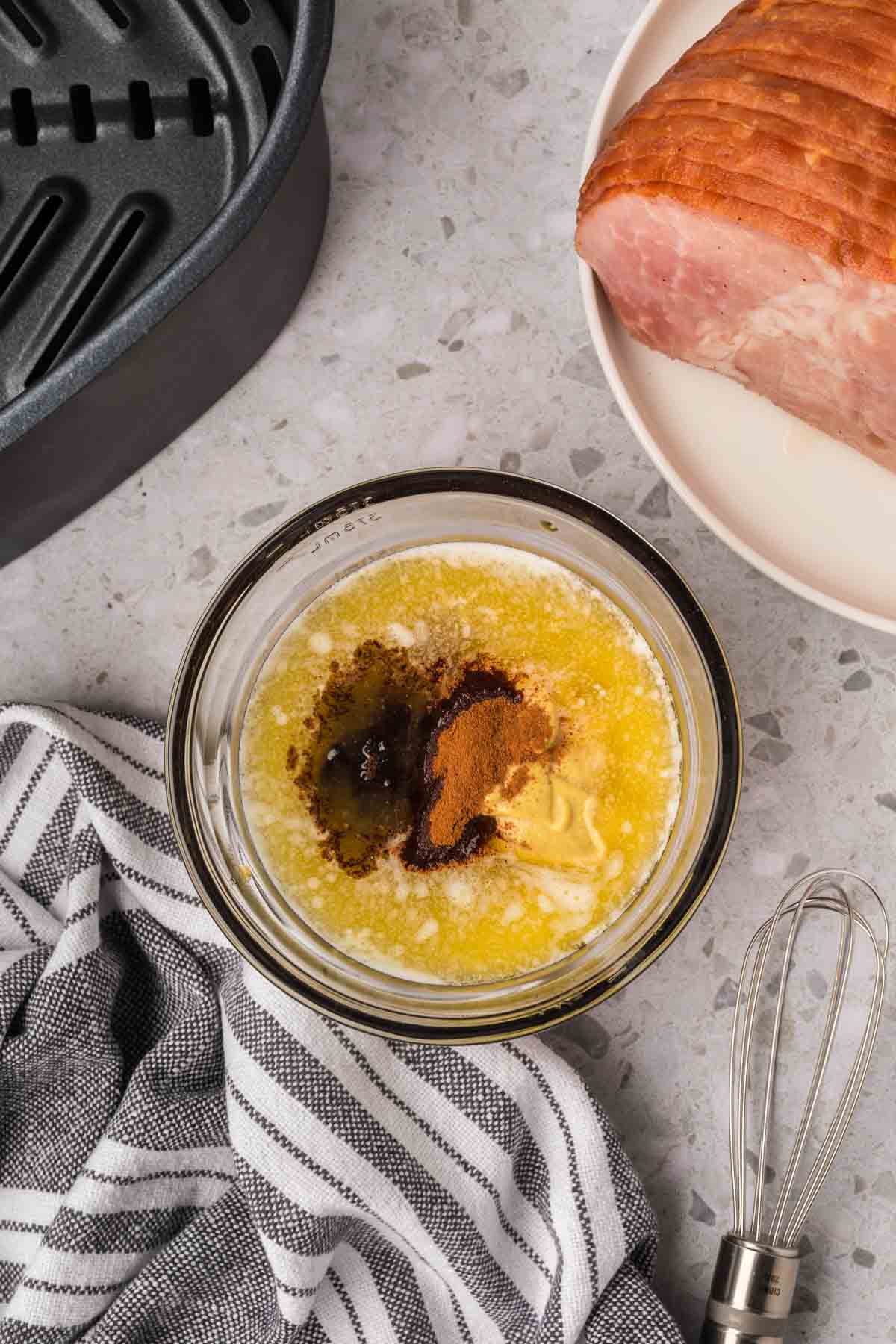 A glass bowl on the countertop holds melted butter with cinnamon and brown sugar. Nearby, an air fryer crisps a spiral-cut ham on a plate, while a whisk, striped cloth, and roasting pan handle lie in view.