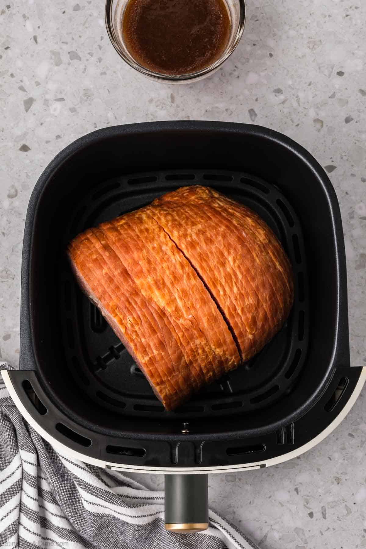 A sliced ham rests inside an air fryer basket on a speckled countertop. Nearby, a bowl of brown glaze awaits, while a striped kitchen towel peeks in from the bottom left.