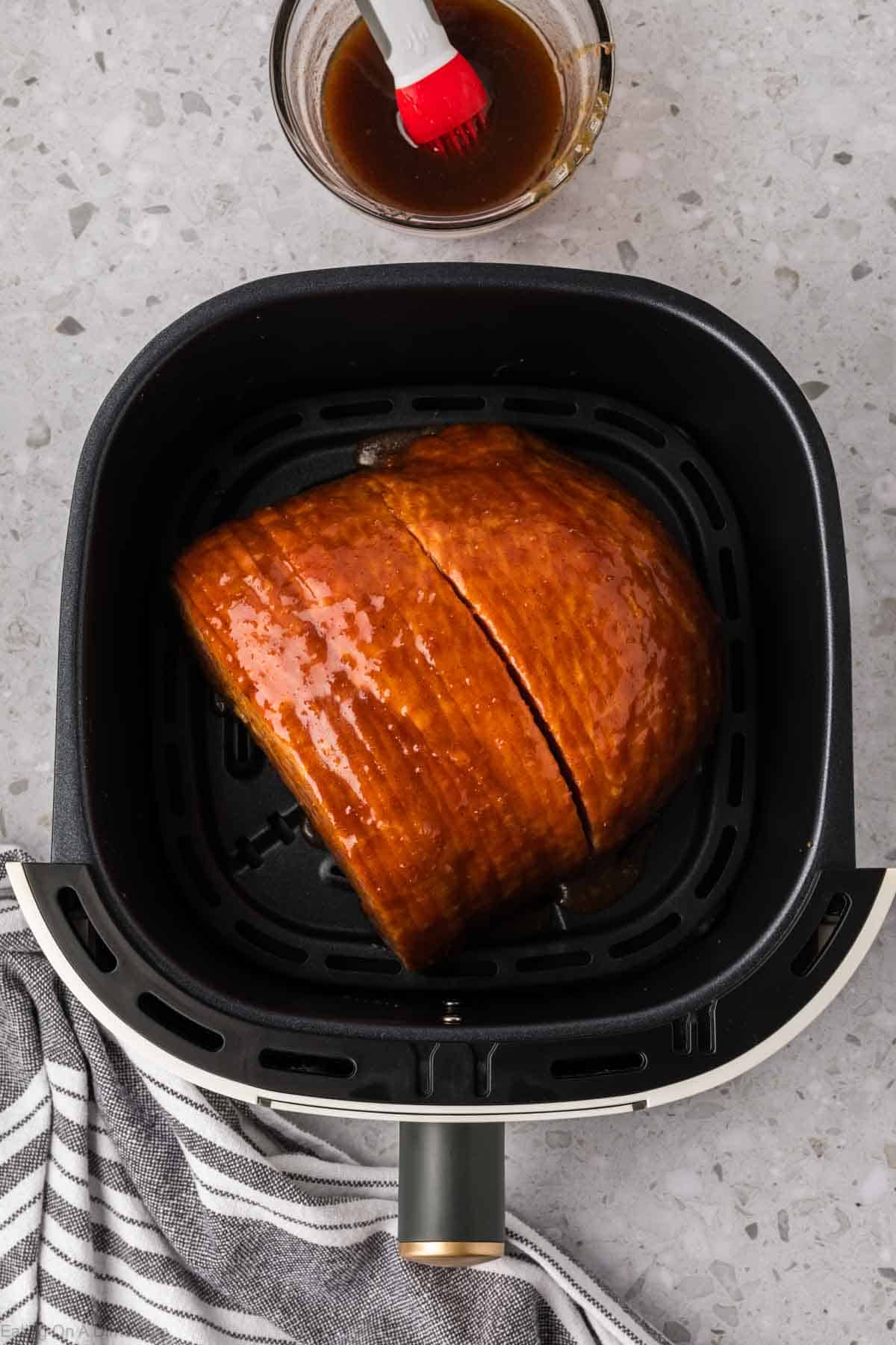 A delicious glazed ham sizzles in the air fryer, with a portion perfectly sliced. A small bowl of glaze and brush sit on the speckled countertop beside it, while a black-and-white striped cloth peeks into view at the bottom of the image.