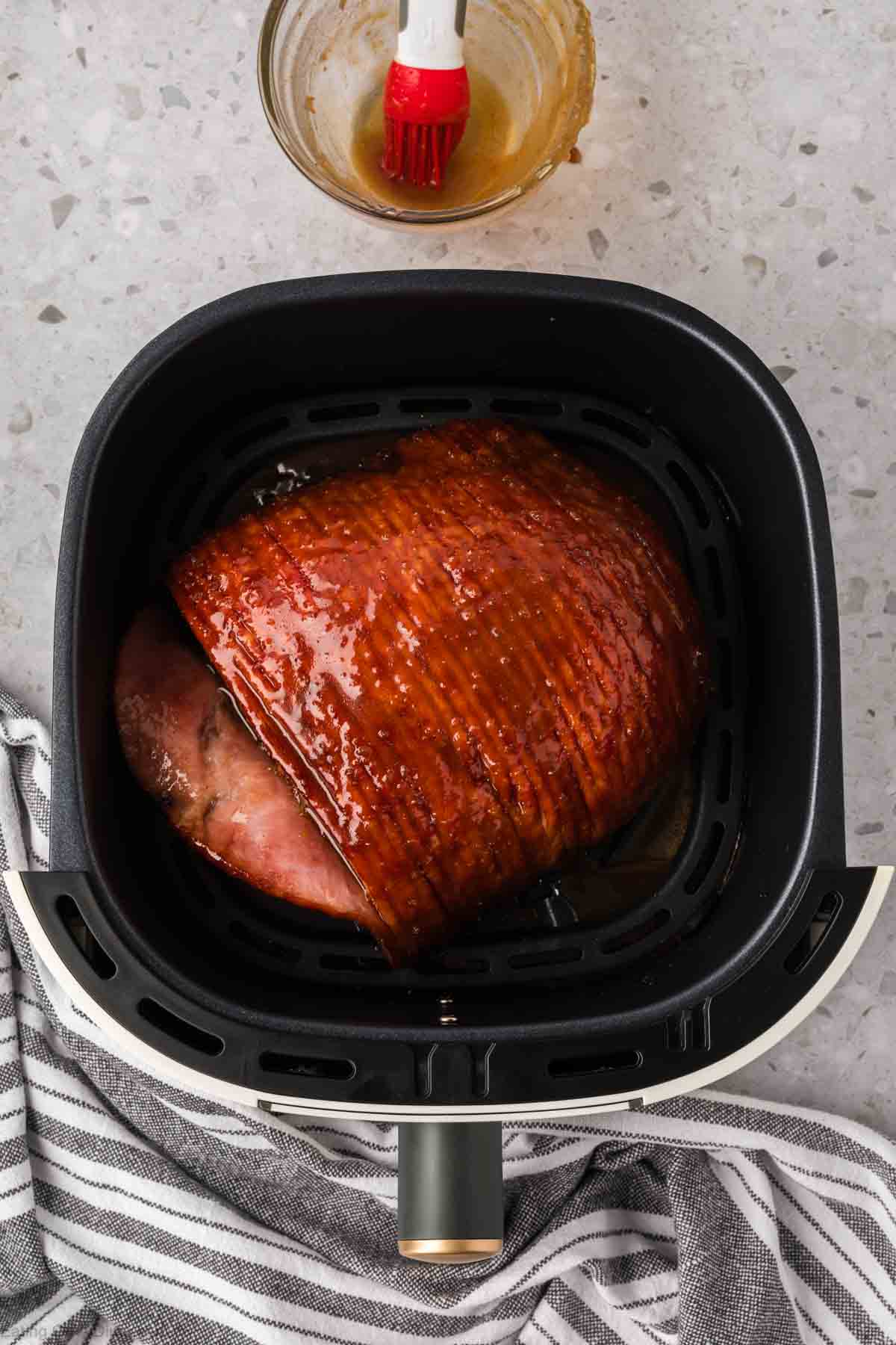 A glazed ham is sizzling in the air fryer basket. A red brush rests inside a glass bowl filled with sauce beside the air fryer on a speckled countertop, with a striped cloth nearby.