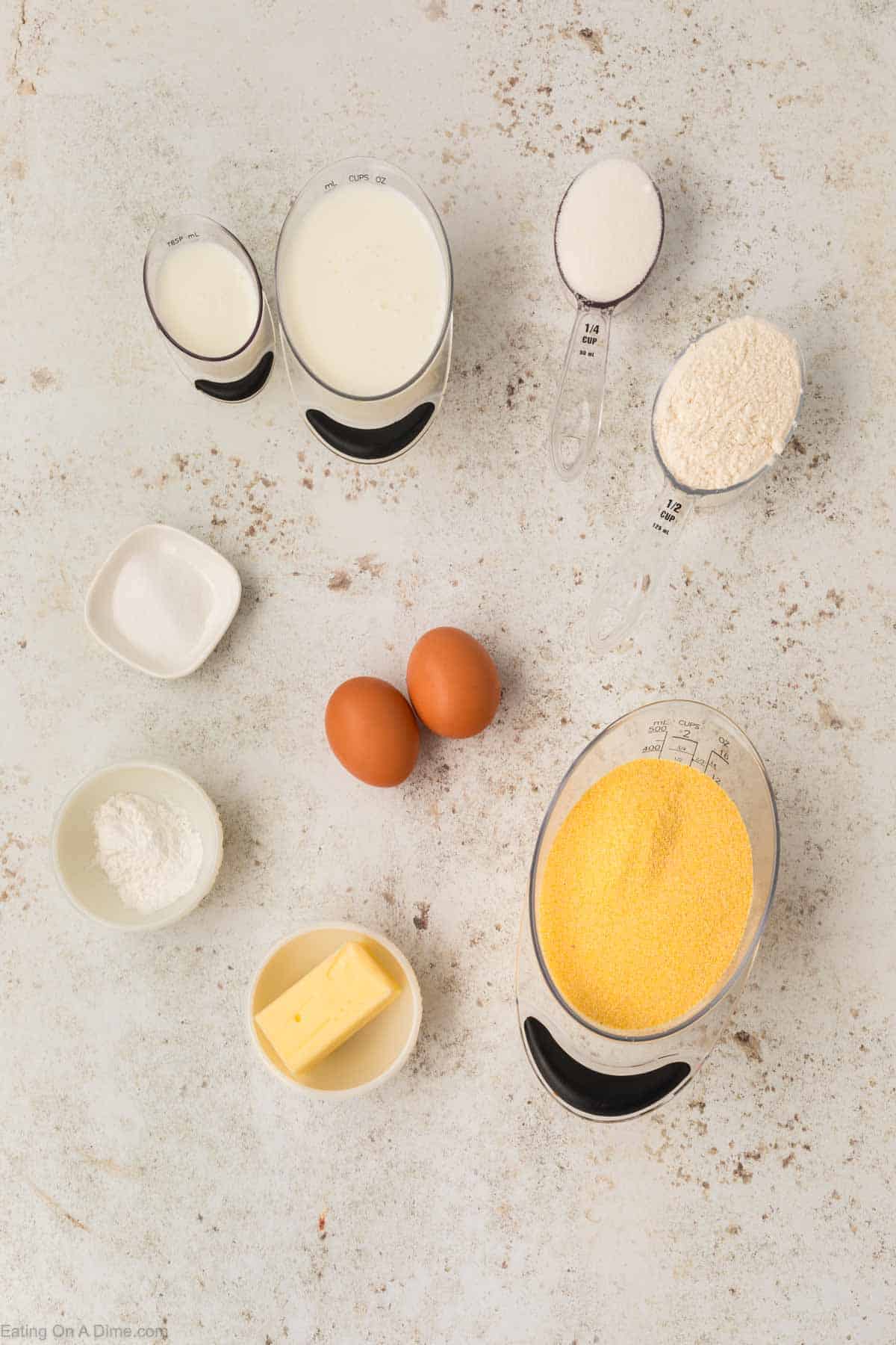 Top-down view of cornbread ingredients on a textured surface, featuring two eggs, a block of butter, and cornmeal. With flour, sugar, baking powder, a small bowl of salt alongside two measuring cups with milk—all ready to be mixed and baked in your trusty cast iron skillet.