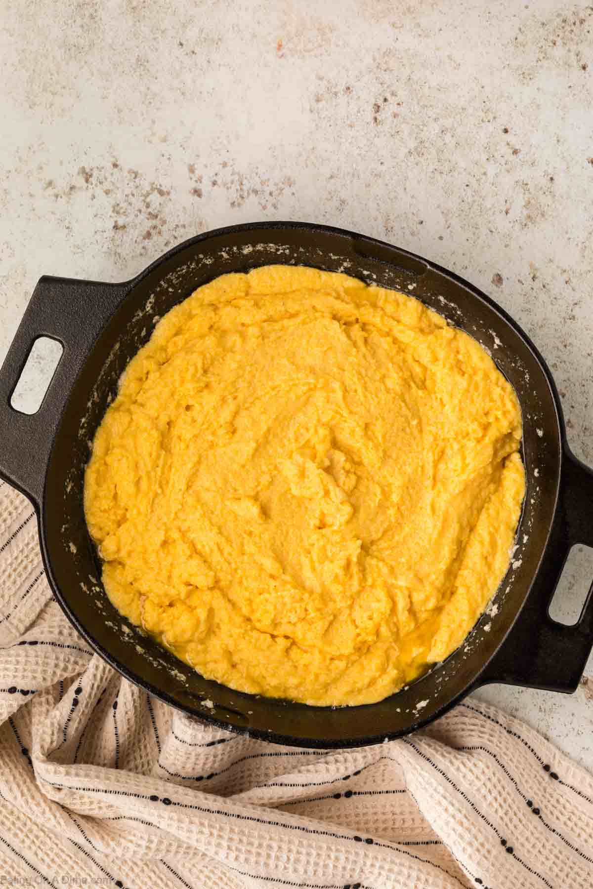 A black cast iron pot, reminiscent of a robust cast iron skillet, is filled with creamy, yellow polenta on a textured surface. A beige and black striped cloth is partially visible underneath the pot.