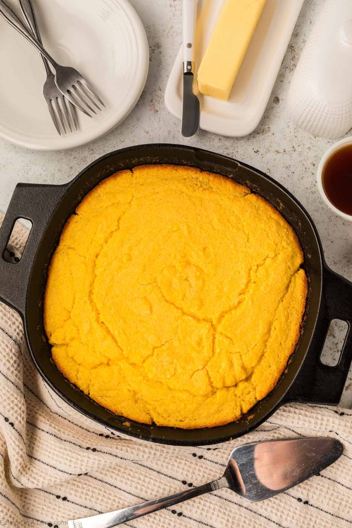 A freshly baked skillet cornbread in a black cast-iron skillet sits on a kitchen counter, surrounded by a butter dish, plates with forks, a cup of coffee, and a striped cloth. A serving spatula is placed next to the pan.