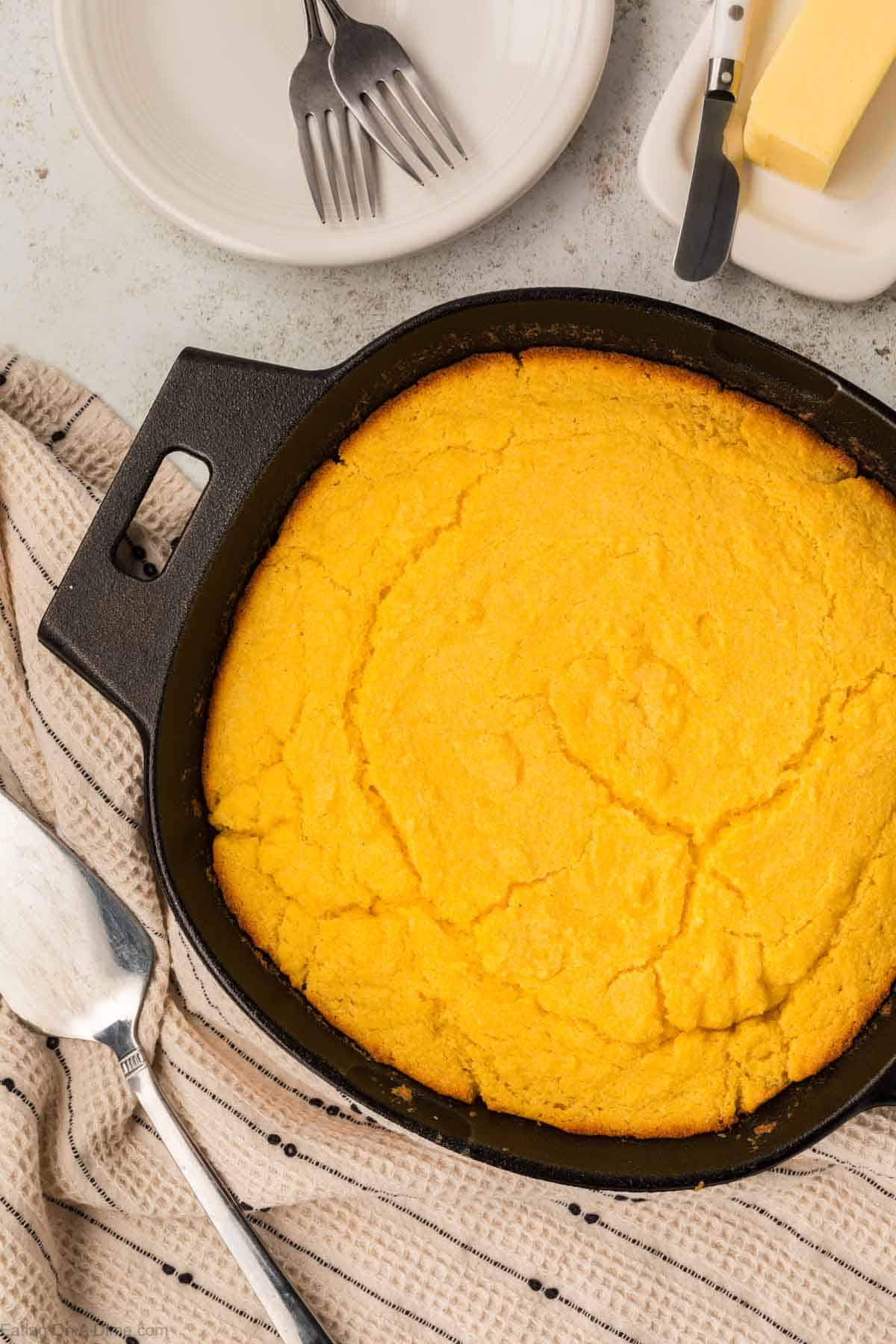 A freshly baked cornbread sits invitingly in a seasoned cast iron skillet on a striped cloth. Nearby are a serving spatula, two forks on a white plate, and a butter dish with a knife, all elegantly arranged on a light-colored surface.