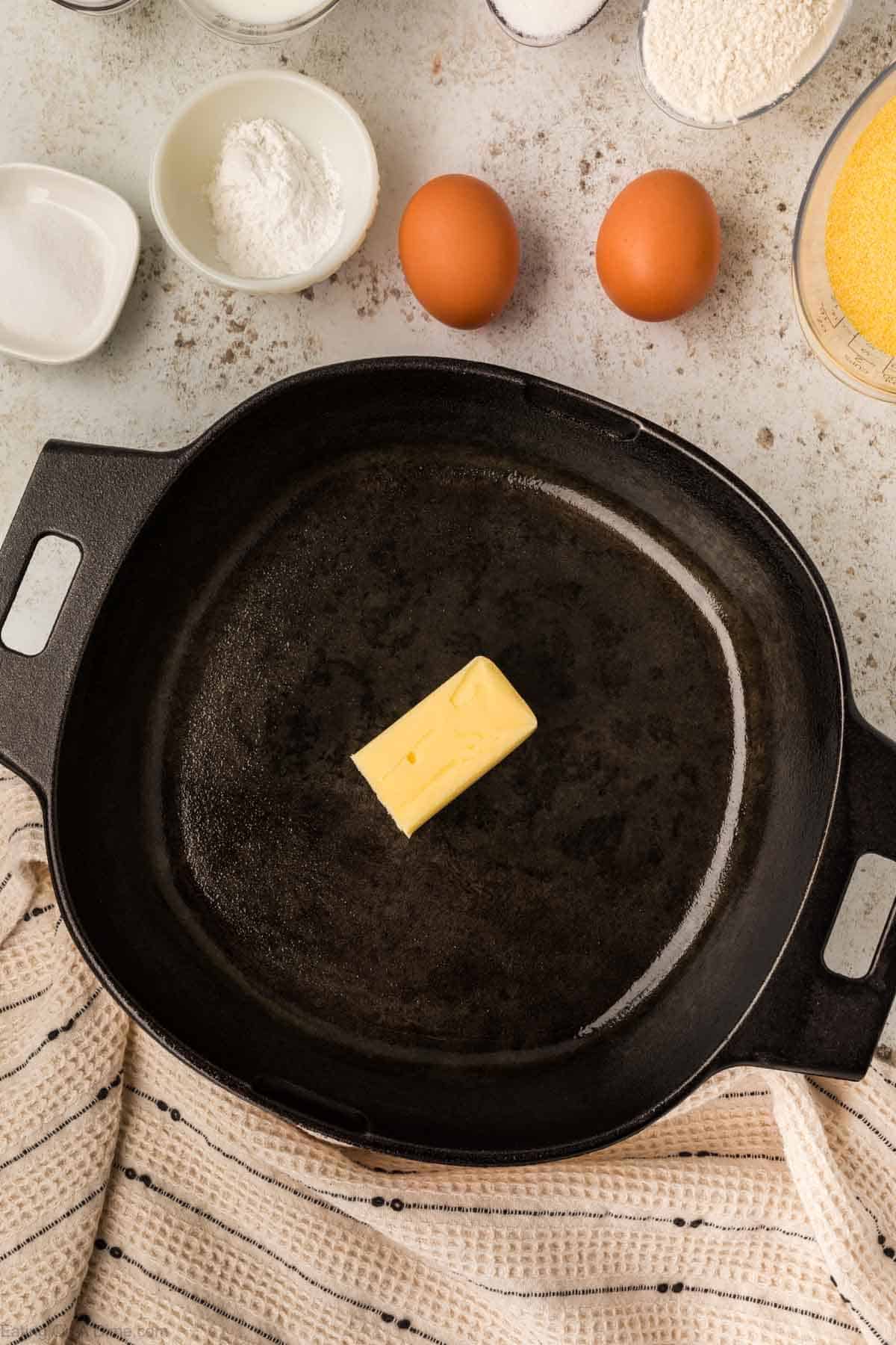 A cast-iron skillet with a stick of butter melting inside sets the stage for perfect cornbread. Surrounding the skillet are two brown eggs, small bowls of dry ingredients, and a measuring cup of cornmeal. A striped cloth lies next to the skillet, completing the cozy kitchen scene.