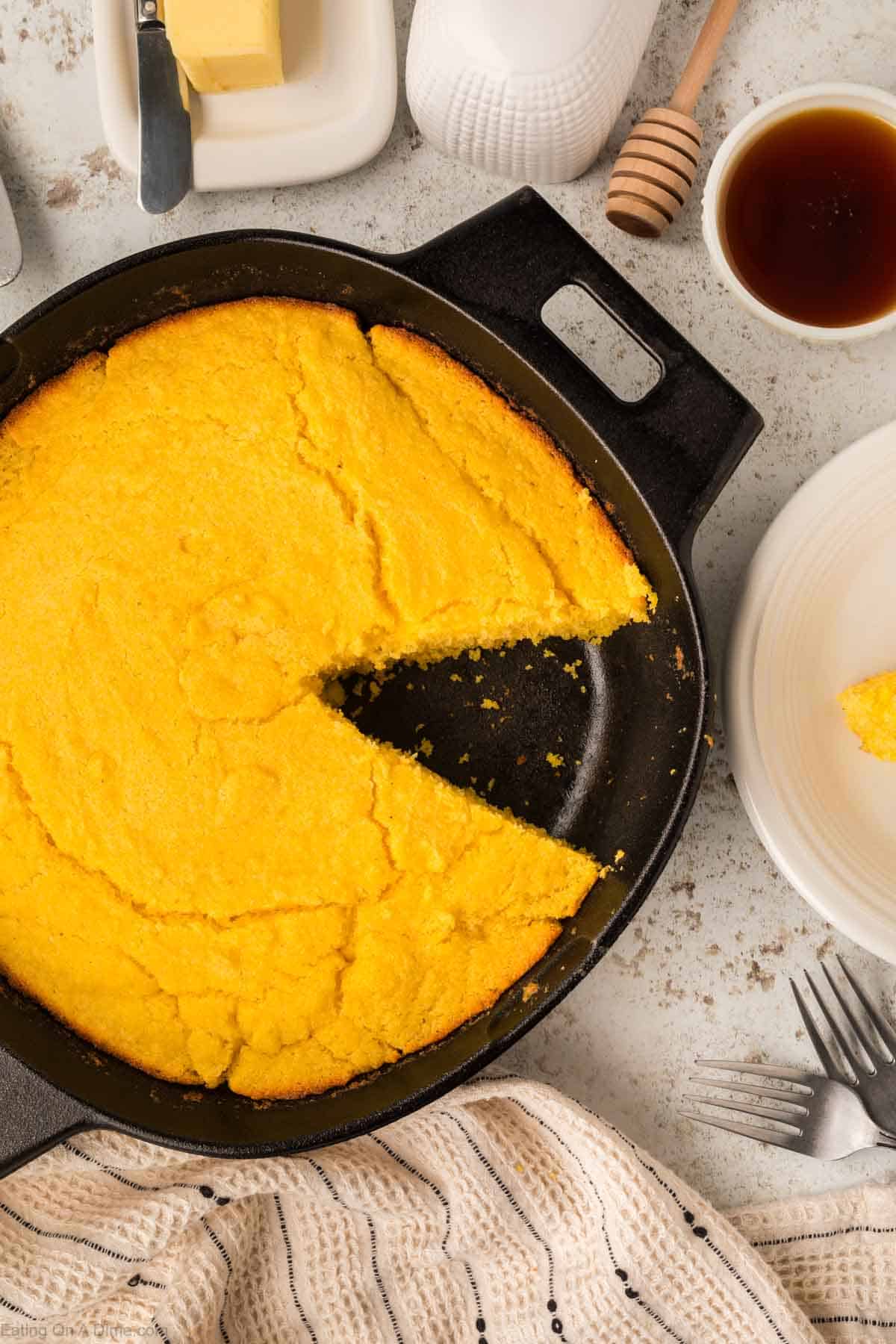 A cast iron skillet with freshly baked cornbread, one wedge cut out, sits next to butter, a honey dipper, and a cup of tea. A plate with a piece of skillet cornbread is on the side, all set on a textured light surface. A striped cloth and fork complete the setting.