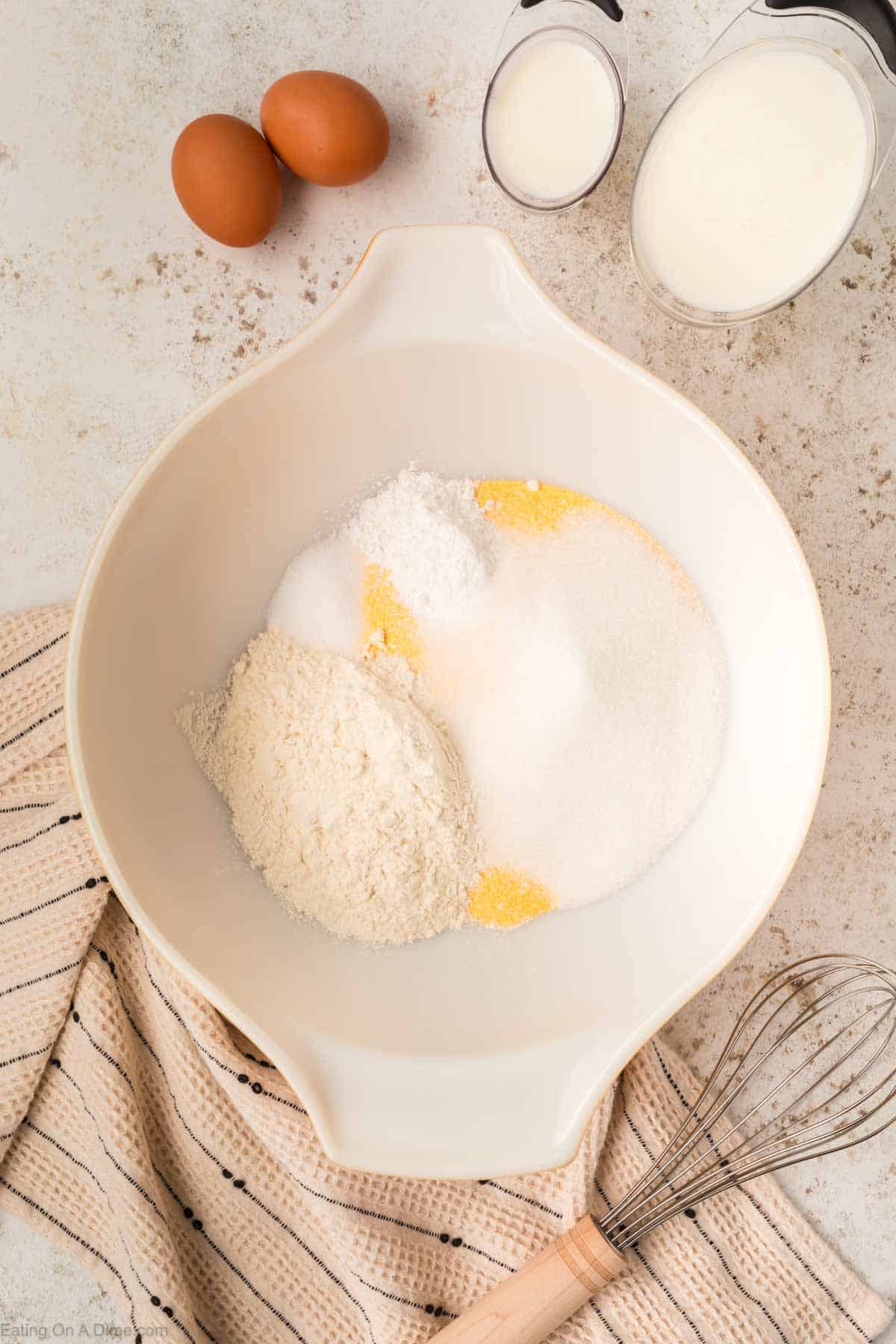 A mixing bowl containing flour, sugar, and other baking ingredients sits on a patterned cloth. Nearby are two eggs, a whisk, a cup of milk, and a cup of oil on a light-colored countertop—perfect for whipping up cornbread for the cast iron skillet.