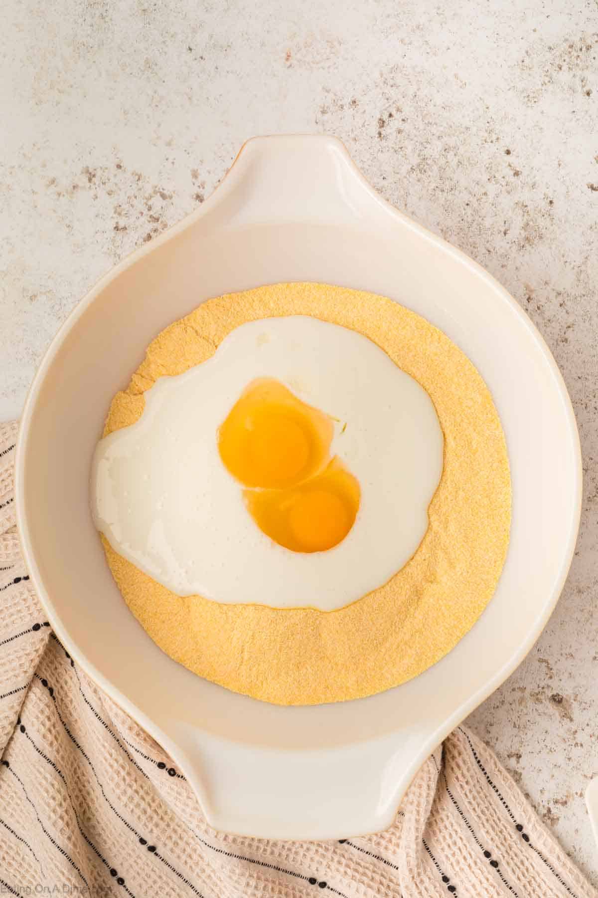 A white mixing bowl containing cornmeal, perfect for cornbread, is topped with milk and two cracked eggs. The bowl rests on a speckled countertop beside a beige striped cloth, ready to be poured into the waiting cast iron skillet.