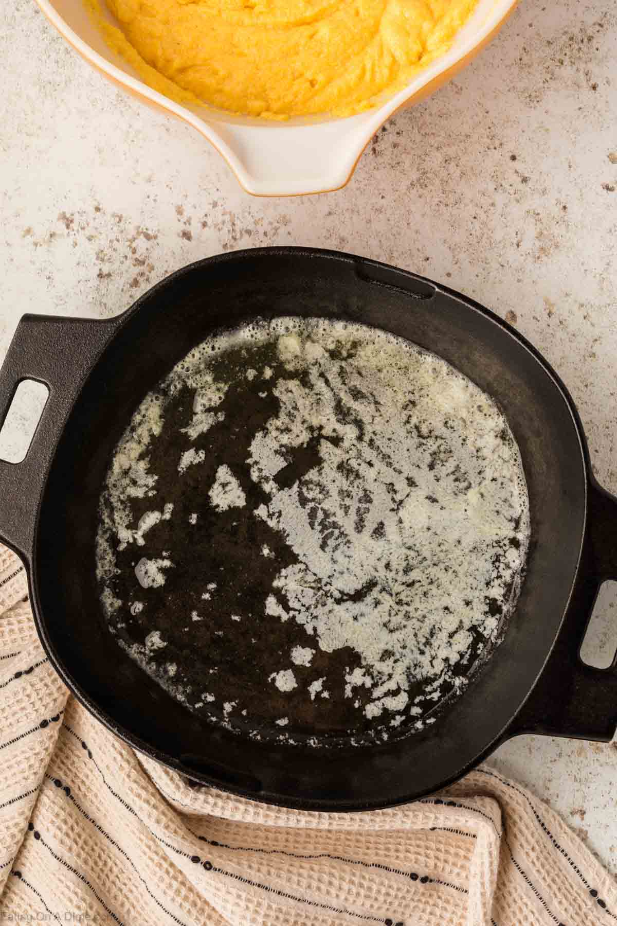 A black cast iron skillet with melted butter sits on a speckled countertop, ready for the cornbread batter waiting in a nearby bowl. A beige striped cloth is draped beside the skillet, adding a touch of rustic charm.