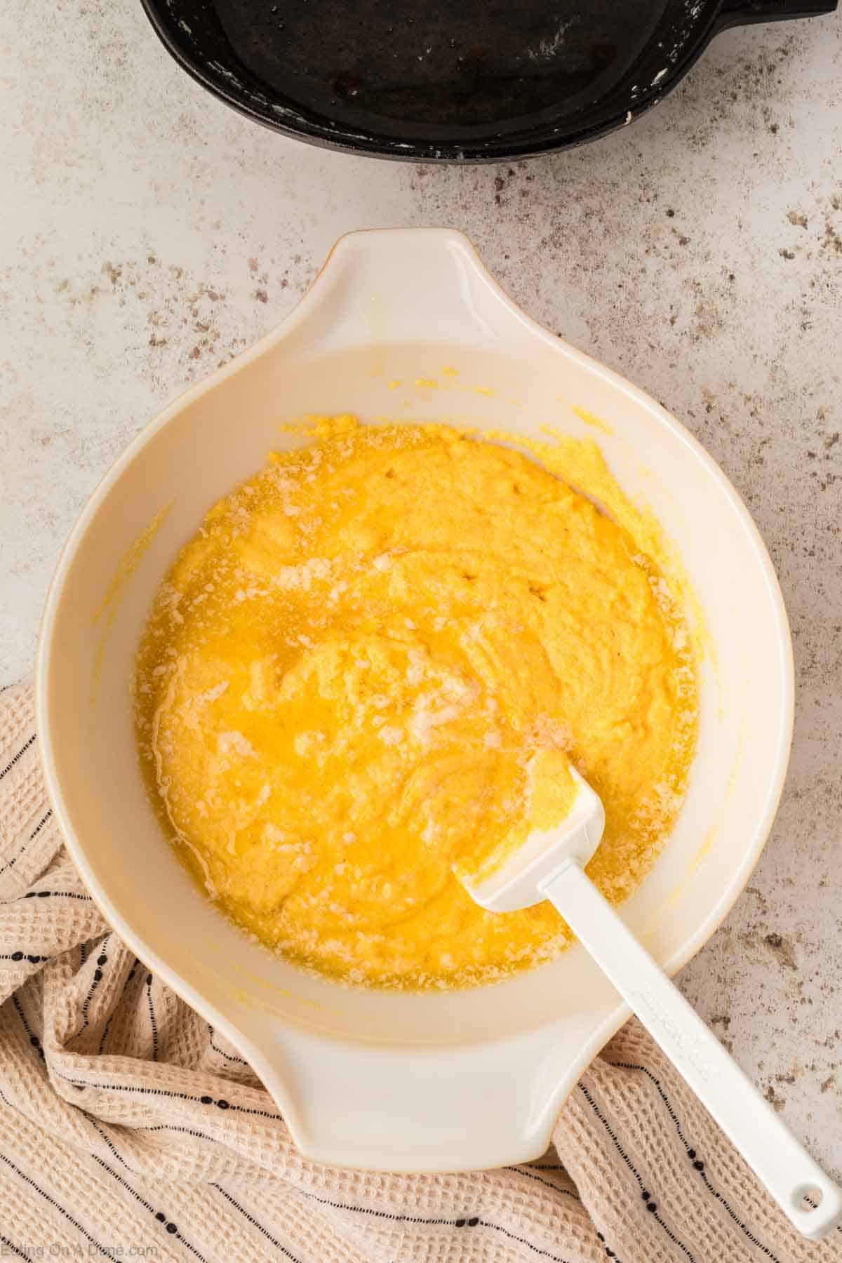 A white bowl contains a mixture of beaten eggs and other ingredients for skillet cornbread, with a white spatula resting inside. The bowl is placed on a gray countertop, and a striped cloth is partially visible beside it.