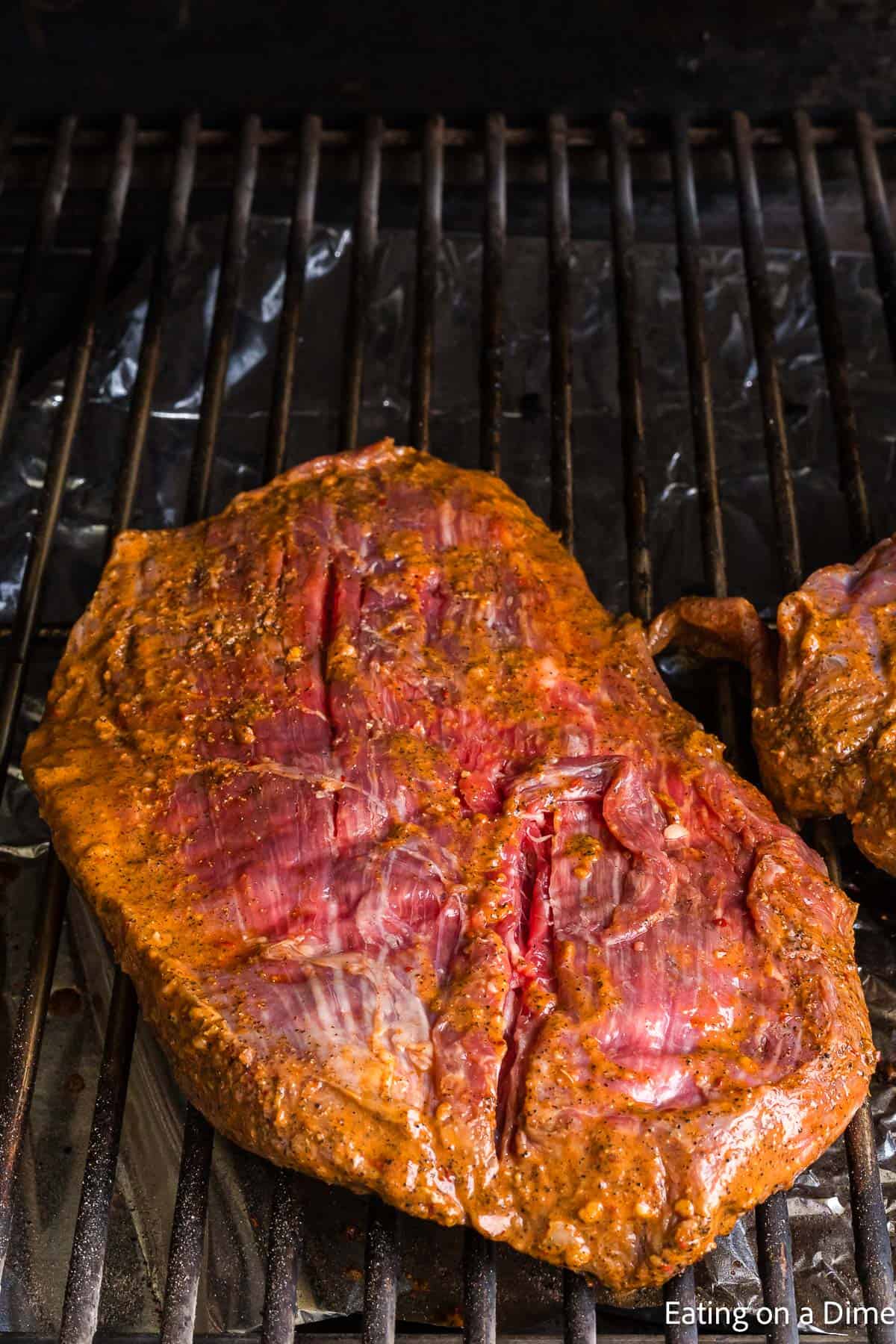 A seasoned piece of meat is cooking on a grill, reminiscent of a Chipotle steak. Marinated with spices, it boasts a reddish-brown appearance, while the grill grates peek out beneath.