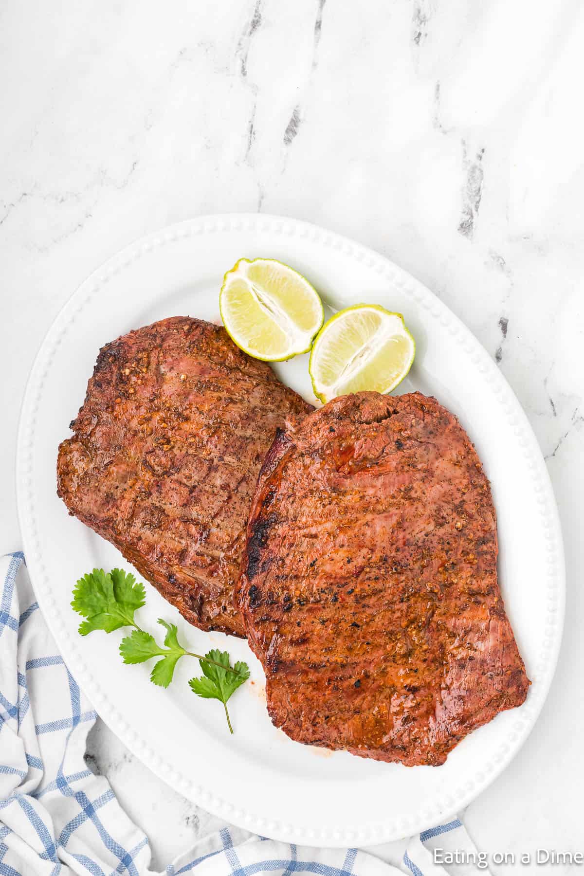 Two grilled steaks on a white oval platter, reminiscent of a Chipotle steak recipe, garnished with fresh cilantro and lime wedges. The platter sits elegantly on a marble surface beside a folded blue and white checkered cloth, inviting you to savor this delicious copycat creation.