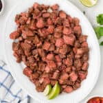 A large white plate is filled with grilled steak cubes, reminiscent of a Chipotle copycat recipe, garnished with lime wedges. A blue and white checkered napkin is partially visible on the left. Fresh cilantro and half a lime are in the background.