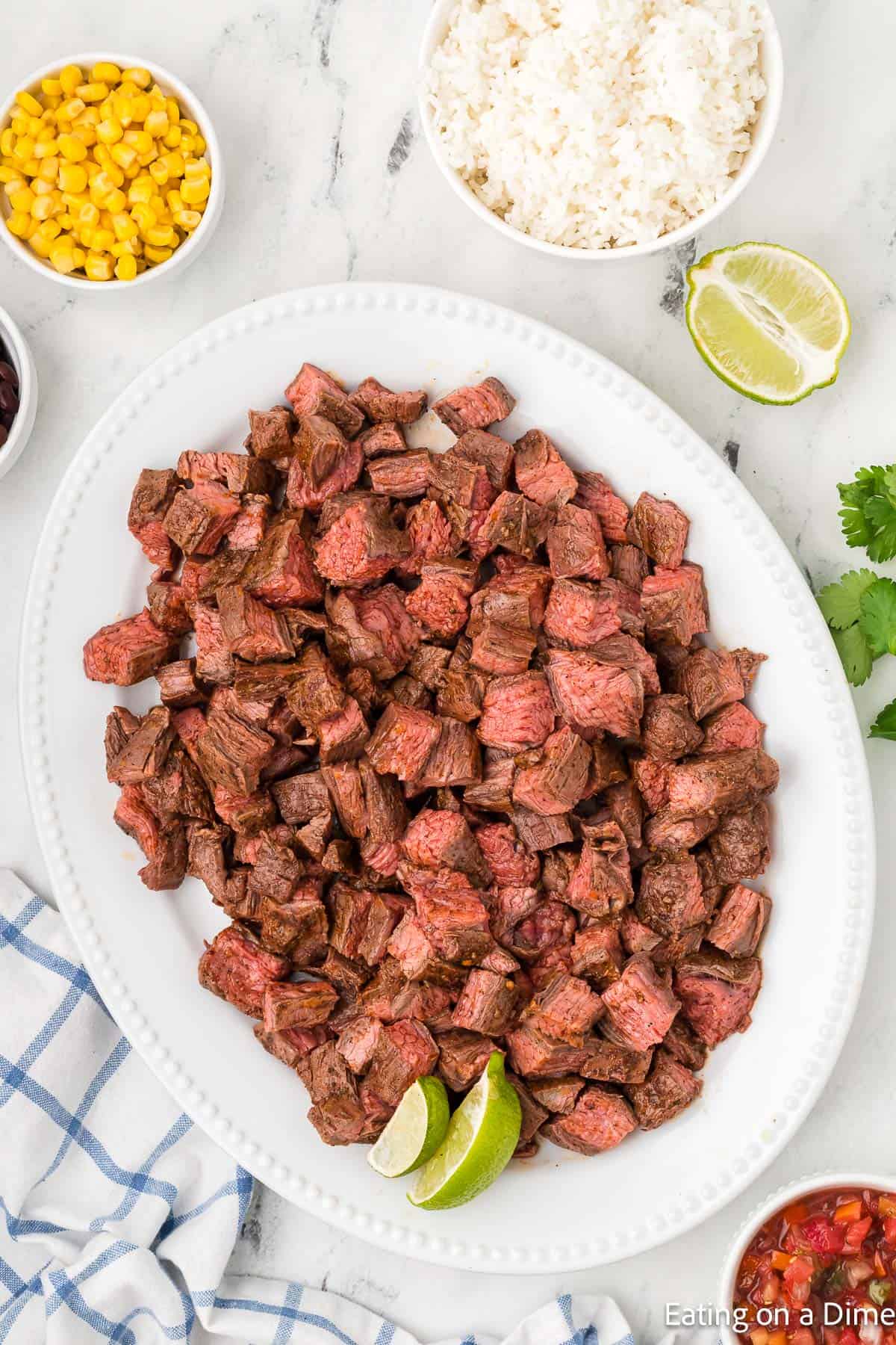 A white platter filled with diced grilled Chipotle steak, garnished with lime wedges. Surrounding the platter are small bowls of corn and rice. A partly visible checkered cloth is in the bottom left corner, while lime halves and cilantro enhance this tempting copycat recipe in the background.