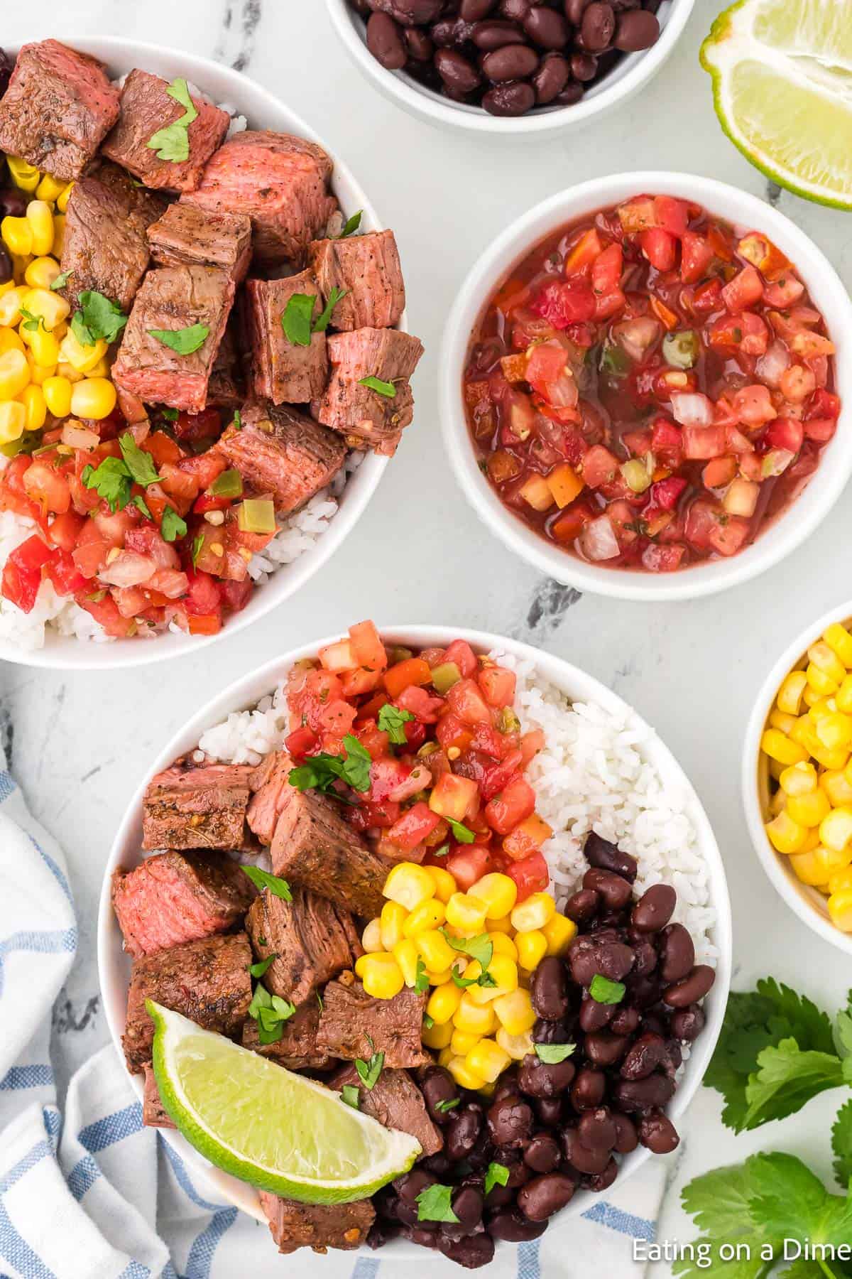 Two bowls brimming with steak, corn, black beans, diced tomatoes, and cilantro over rice, accompanied by lime wedges. Channeling a Chipotle steak vibe, additional bowls of black beans, salsa, sliced lime, and cilantro complete the scene on a striped cloth napkin.