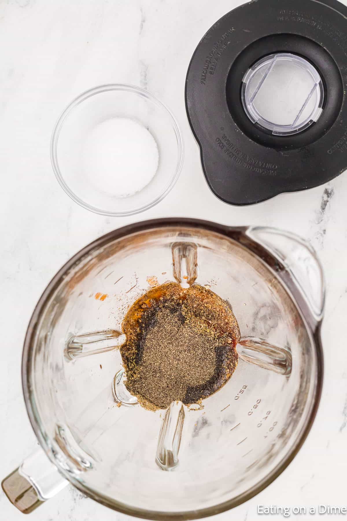 A blender with various spices sits on a marble countertop, ready to create a copycat recipe of Chipotle steak. Nearby, there's a clear bowl of salt and the black blender lid, eagerly awaiting the flavorful adventure.