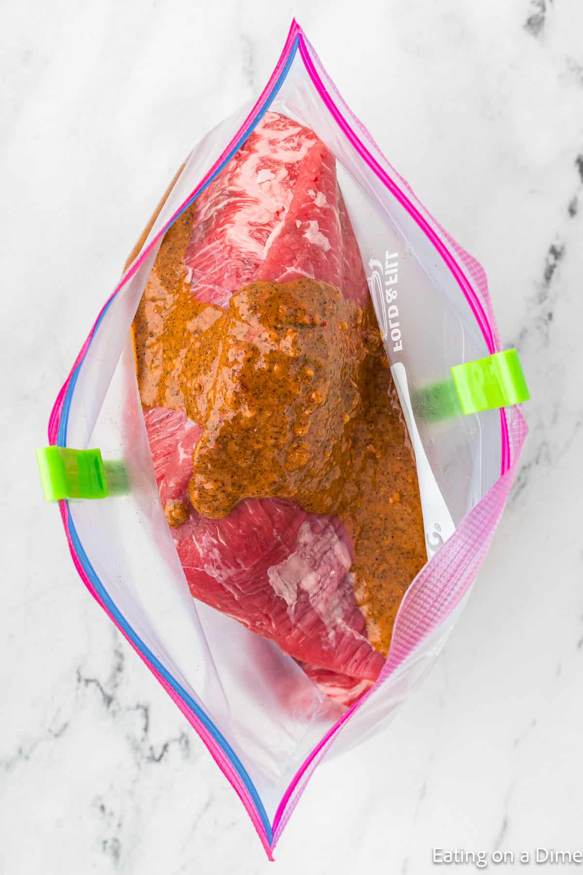 Aerial view of a piece of steak marinating in a clear ziplock bag filled with a chipotle-inspired reddish-brown marinade, secured with green bag clips. The copycat creation rests on a marble surface, promising bold flavors.