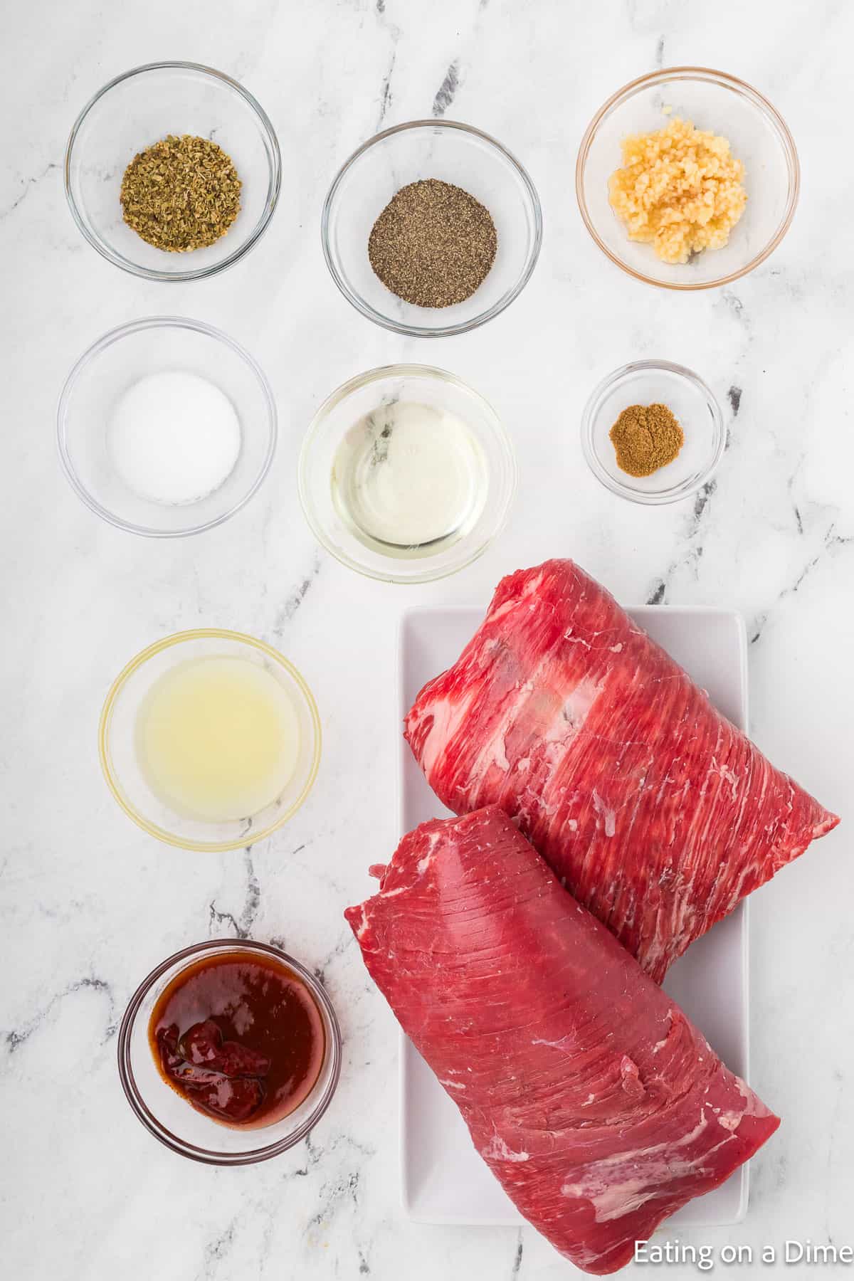 Raw beef on a plate with small bowls of spices and ingredients, including minced garlic, ground black pepper, oregano, salt, lime juice, and chipotle sauce as part of a copycat Chipotle steak recipe arranged on a marble surface.