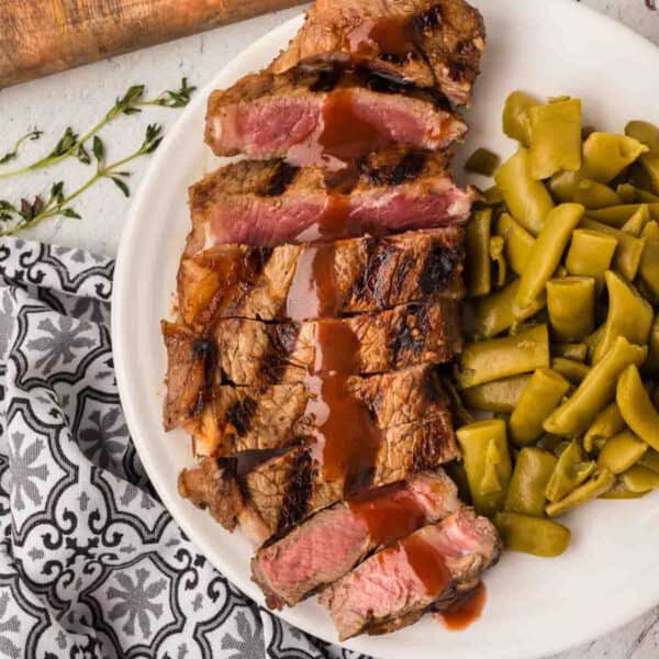 A plate with sliced grilled steak drizzled with a rich red wine steak sauce, served alongside cooked green beans. A patterned black and white napkin is partially visible next to the plate.