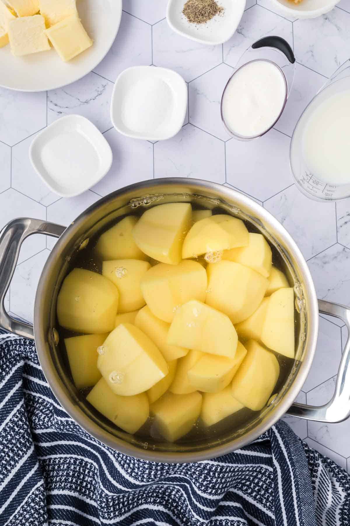 Diced potatoes in a large pot of water with a plate of butter slices, small bowl of salt, pepper, and heavy cream