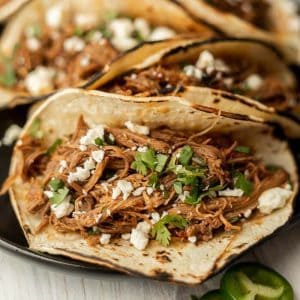 Close-up of a delicious taco, part of Meal Plan 32, filled with shredded meat, crumbled cheese, and fresh cilantro on a lightly charred tortilla. Additional tacos are visible in the background on a sleek black plate.