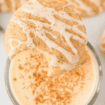 A cinnamon-flavored cookie with white icing drizzle sits on the edge of a glass filled with creamy eggnog, topped with a sprinkle of cinnamon—a perfect treat for those exploring recipes with eggnog. Another cookie is partially visible in the background.