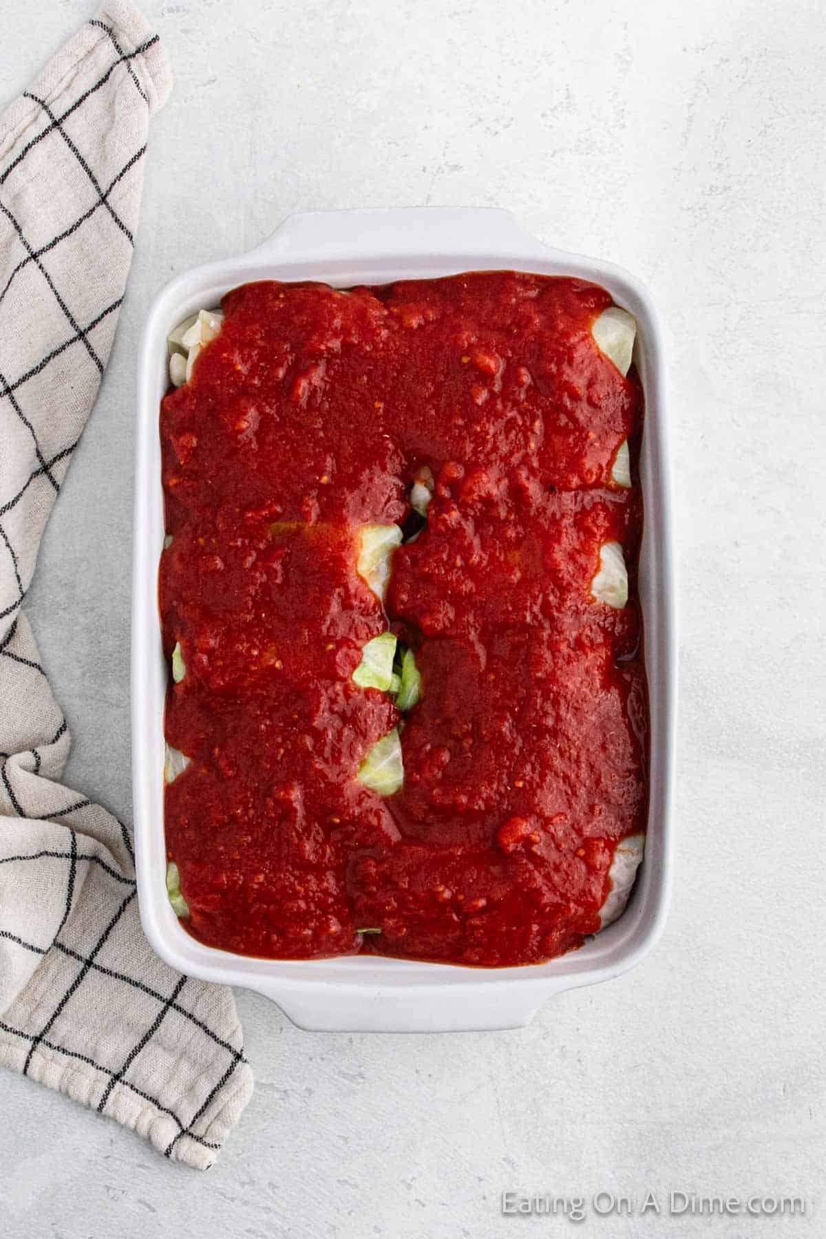 A white baking dish filled with flavorful cabbage rolls smothered in tomato sauce sits on a light grey surface, next to a black and white checkered cloth. This inviting recipe is a staple of classic cuisine.