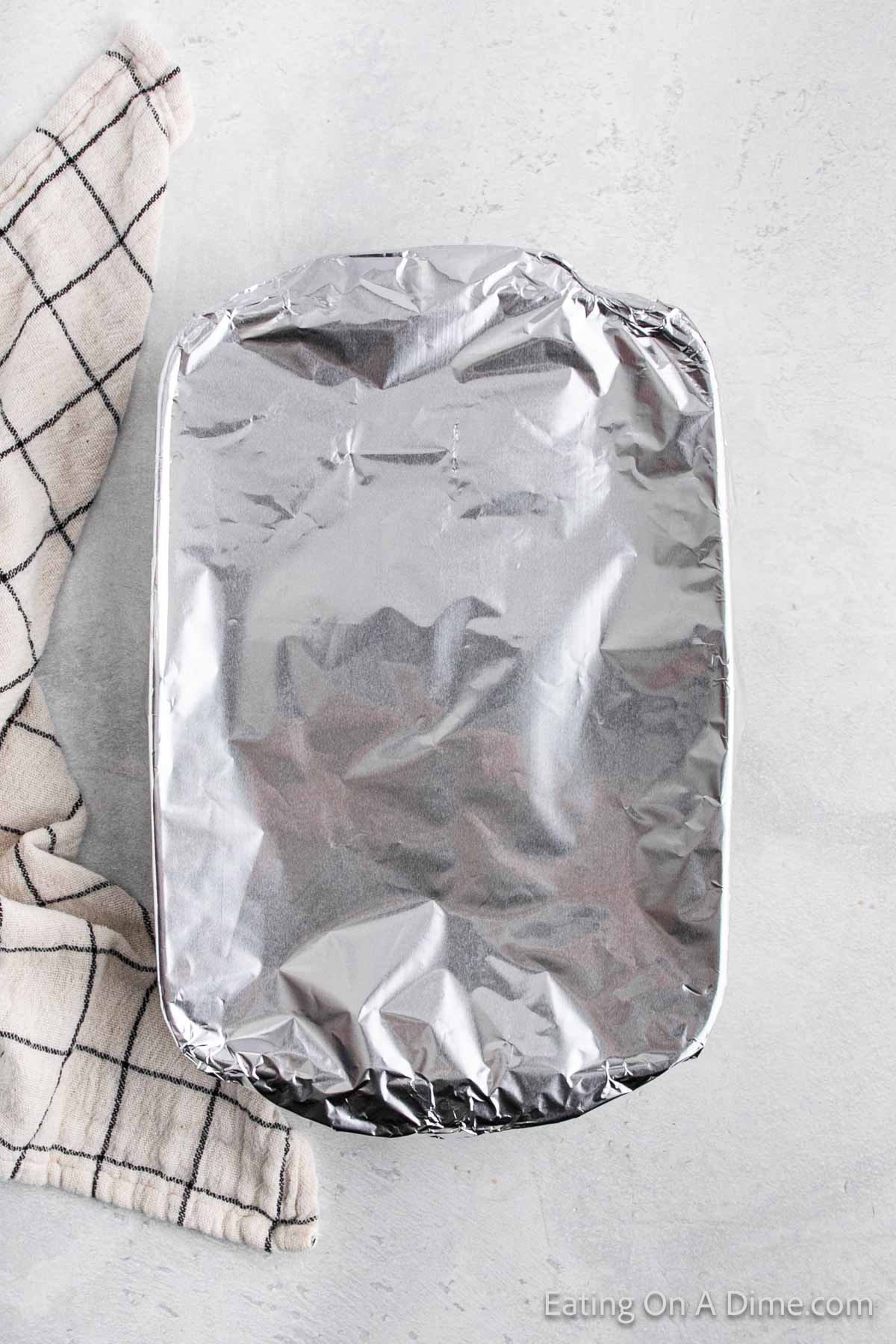 A rectangular baking dish covered with shiny aluminum foil sits on a light gray surface, hiding delicious cabbage rolls. To the left, a black and white checkered cloth adds a touch of charm.