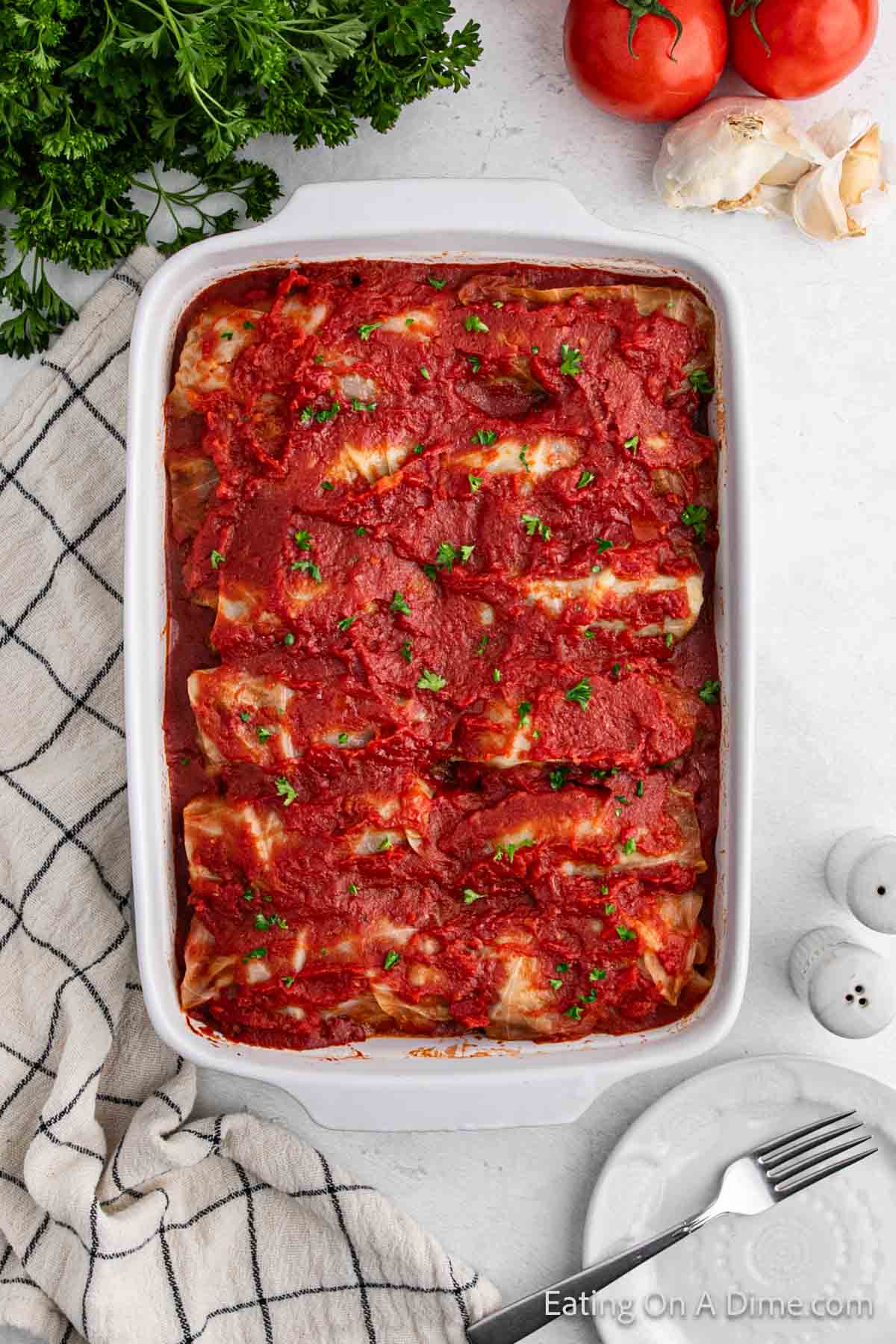 A baking dish filled with manicotti covered in marinara sauce and garnished with chopped herbs, reminiscent of hearty cabbage rolls. The dish is set on a checkered cloth with a fork and spoon beside it. Fresh parsley and tomatoes are placed in the background.