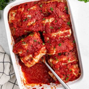 A white baking dish showcases savory cabbage rolls engulfed in a rich tomato sauce, garnished with chopped parsley. A spoon is placed on the side, ready for serving. A striped kitchen towel is partially visible to the left.