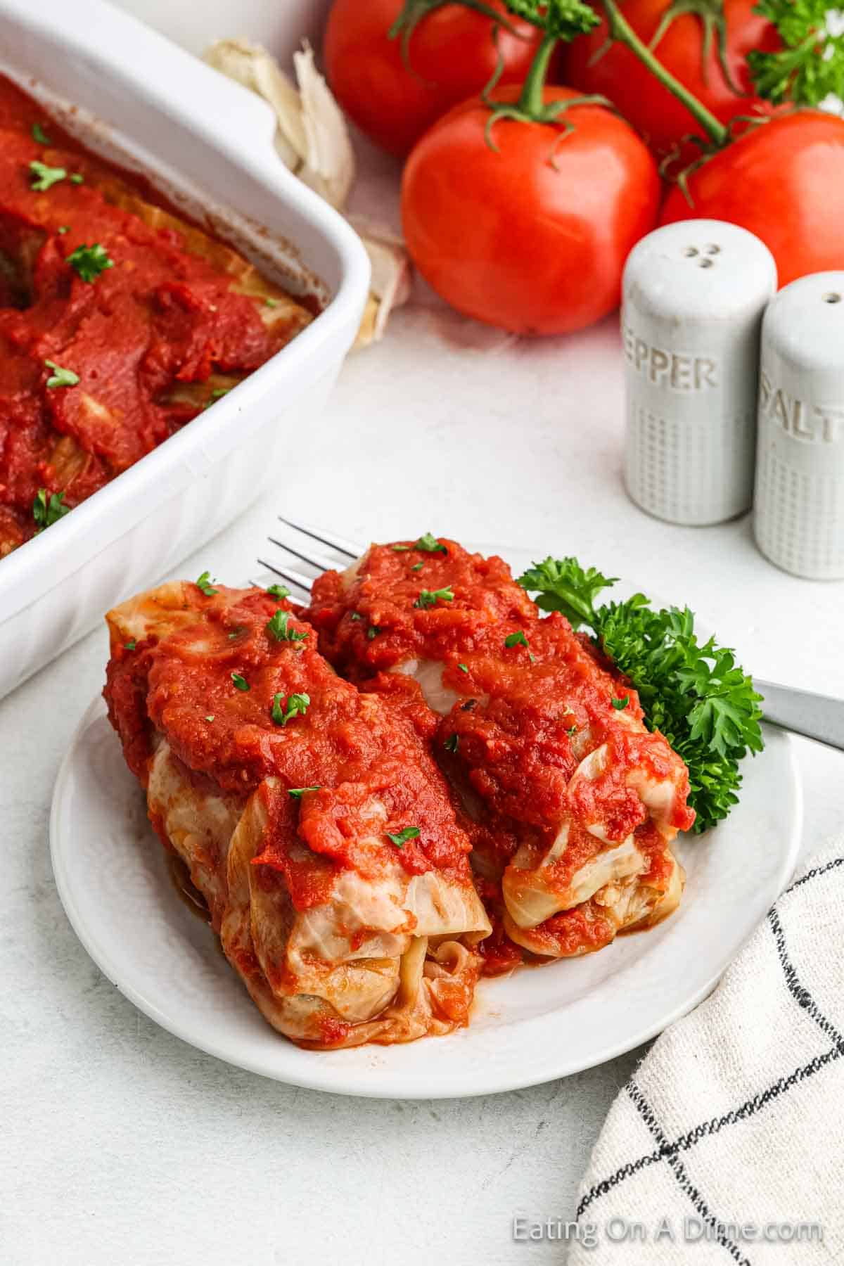 Two cabbage rolls topped with tomato sauce sit on a plate. Garnished with parsley, these delicious creations are a staple in any cooking recipe book. Accompanied by a fork, they share the scene with fresh tomatoes, more cabbage rolls, a towel, and salt and pepper shakers in the background.