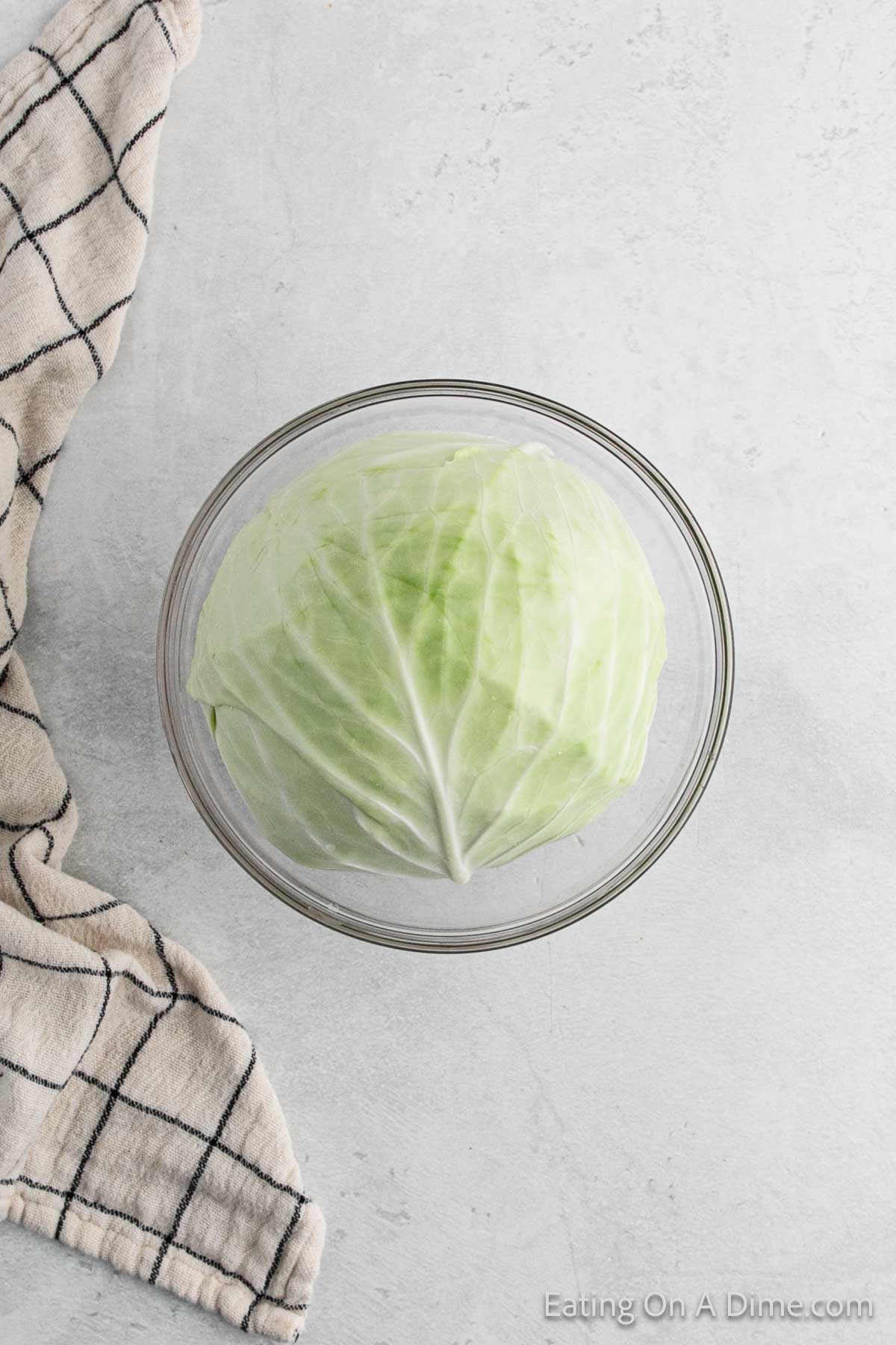 A whole head of cabbage, ideal for making savory cabbage rolls, sits in a glass bowl on a light gray surface. To the left, a checkered cloth is partially visible.