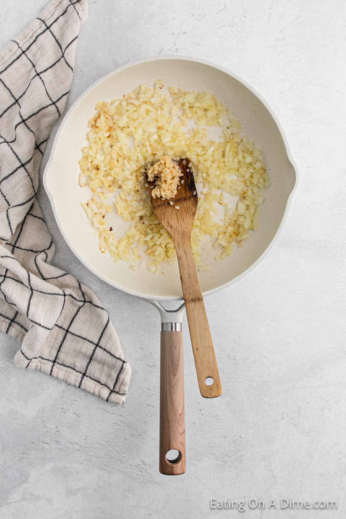 A white skillet on a gray surface contains sautéed onions and cabbage, with a wooden spoon resting inside. A checked kitchen towel is placed nearby, suggesting the start of a cozy recipe.