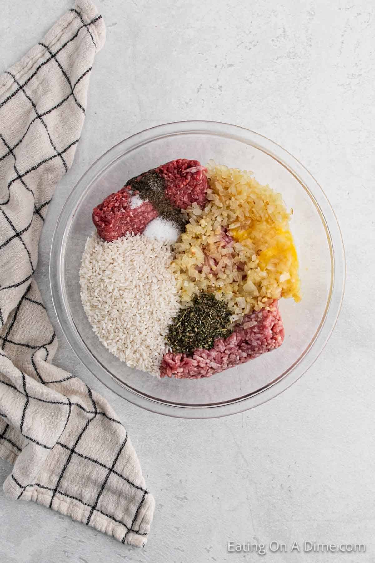 A glass bowl on the countertop holds the beginnings of savory cabbage rolls: raw ground meat, chopped onions, uncooked rice, and various spices. A checkered kitchen towel lies beside the bowl.