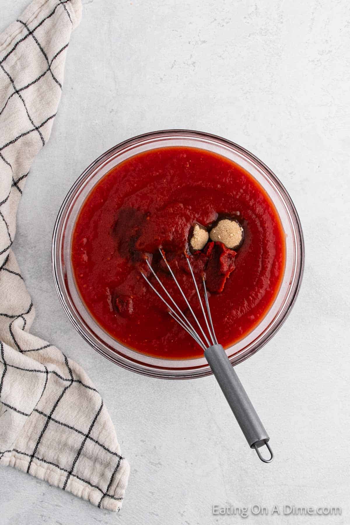 A glass bowl filled with red barbecue sauce mixture, perfect for drizzling over cabbage rolls, contains spices and a whisk. It sits on a light gray surface, while a black and white checkered towel is partially visible to the left of the bowl.