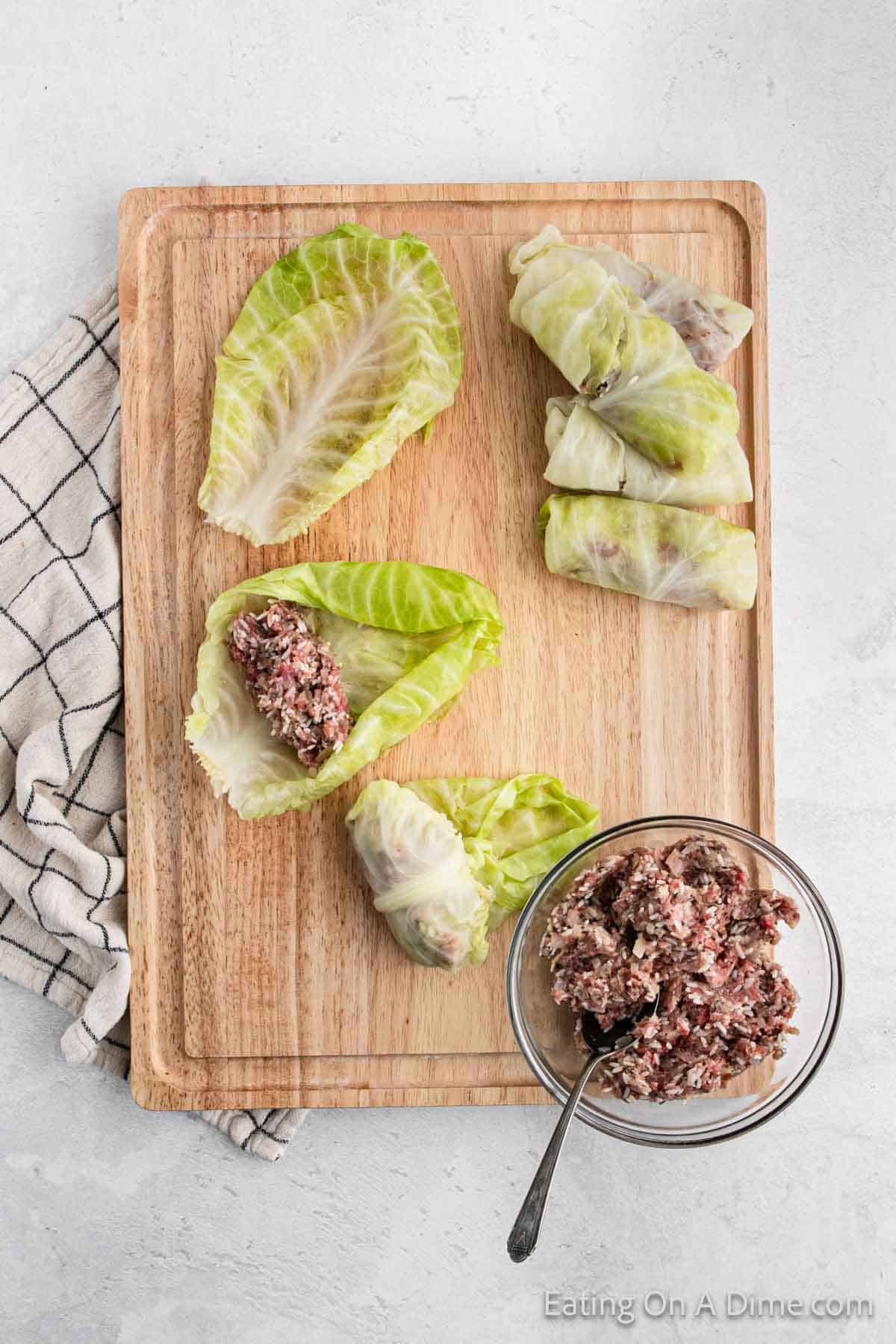 Cabbage leaves are filled with a minced meat mixture on a wooden cutting board, transforming into delicious cabbage rolls. Some rolls are already wrapped neatly. Nearby, a bowl brims with more filling and a spoon. A checkered cloth adds charm to the scene.