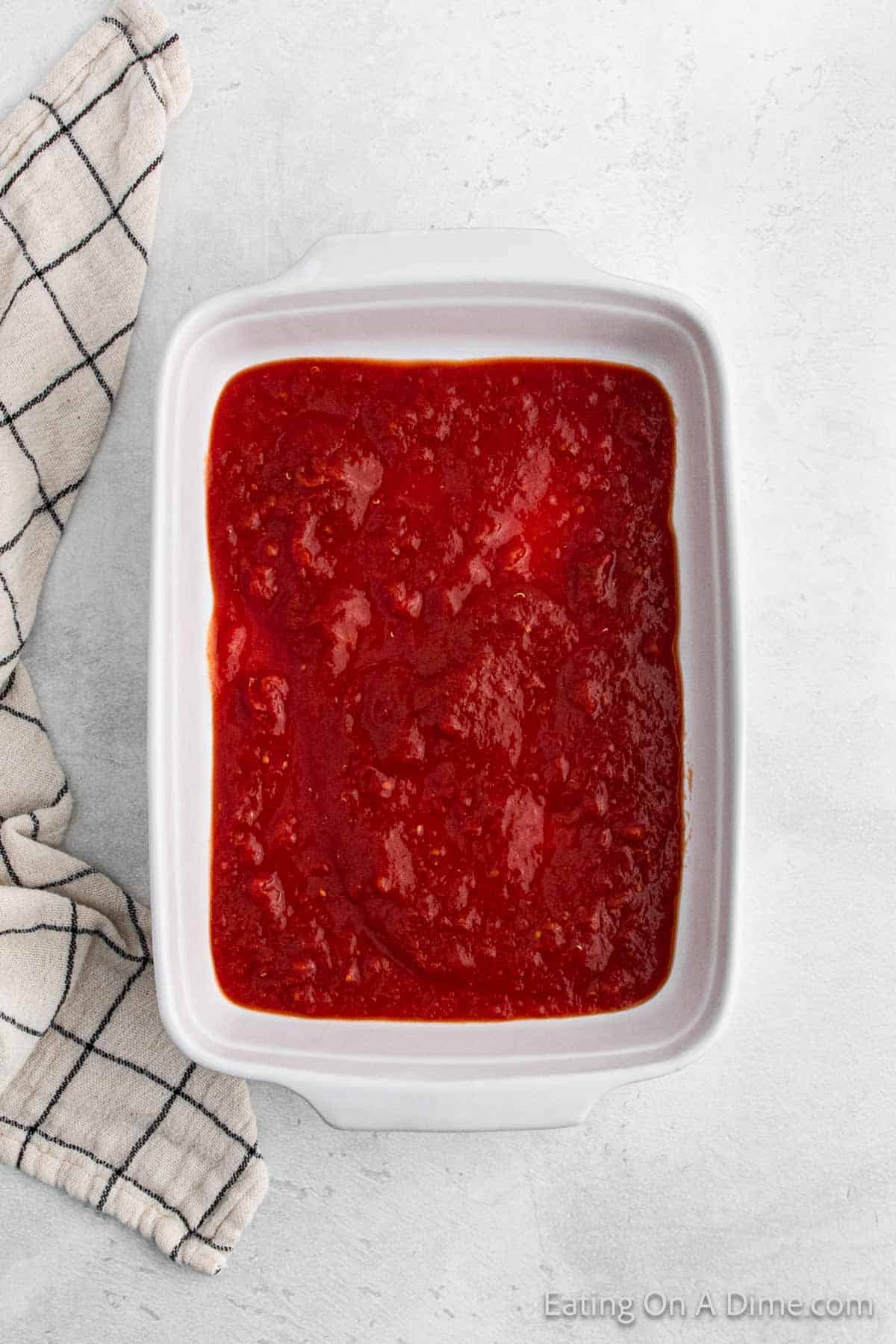 A white rectangular dish filled with smooth red tomato sauce cradles perfectly cooked cabbage rolls. A black and white checkered cloth is partially visible on the left side, all set against a light gray surface in the background.