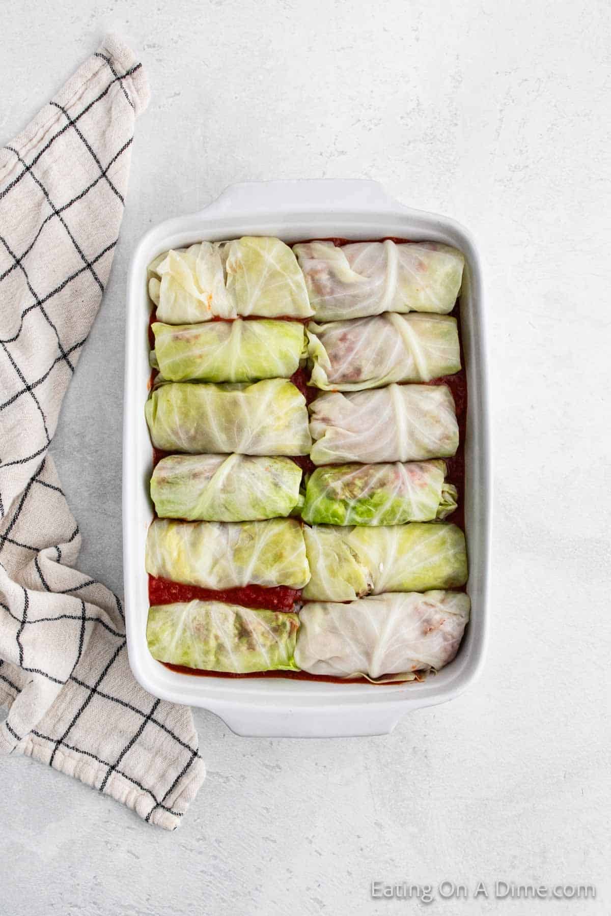 A baking dish filled with neatly arranged cabbage rolls on a light gray surface. The delicious rolls are part of a classic recipe, nestled over a layer of red sauce. Beside the dish, a cloth with a black grid pattern completes the inviting cooking scene.