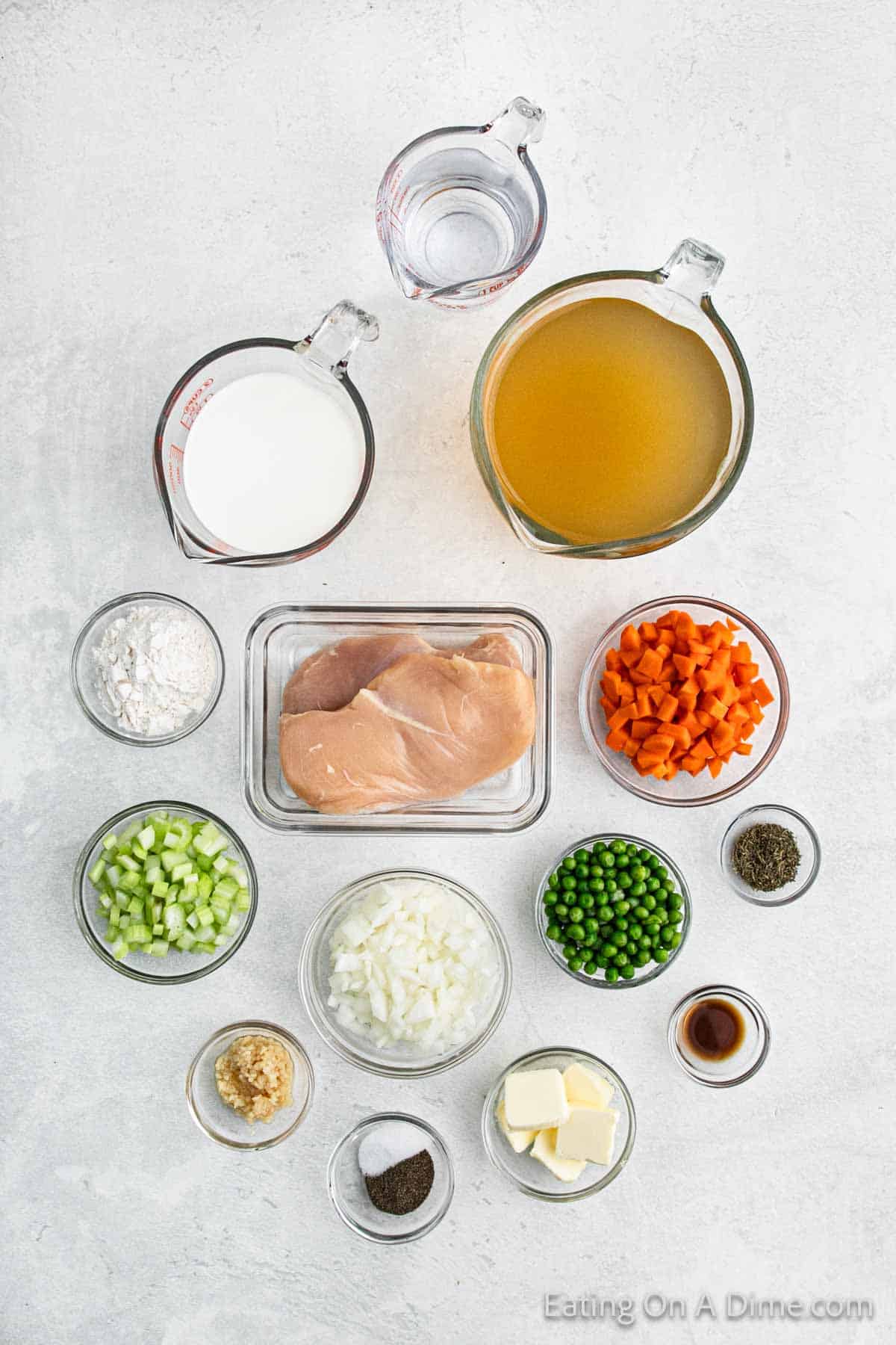 Ingredients for a comforting chicken pot pie soup recipe, reminiscent of chicken and dumplings, include tender chicken breasts, chopped carrots, peas, celery, onions, garlic, butter, flour, milk, chicken broth, water, and seasonings—all arranged in measuring cups and bowls on a light surface.