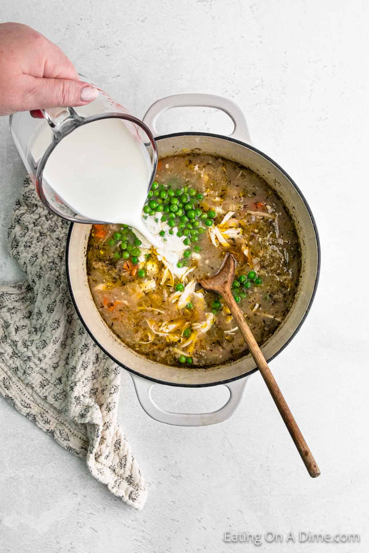 A hand pours milk from a measuring cup into a pot of soup enriched with dumplings. The soup contains peas, shredded chicken, and vegetables. A wooden spoon rests inside the pot, and a patterned cloth is nearby.