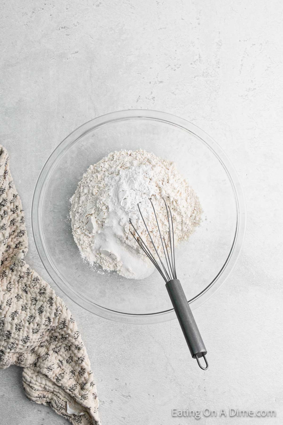 A glass bowl with flour and baking powder being mixed with a whisk, setting the stage for delicious chicken dumplings. A patterned cloth is positioned to the left on a light gray surface, adding a touch of charm to the culinary scene.