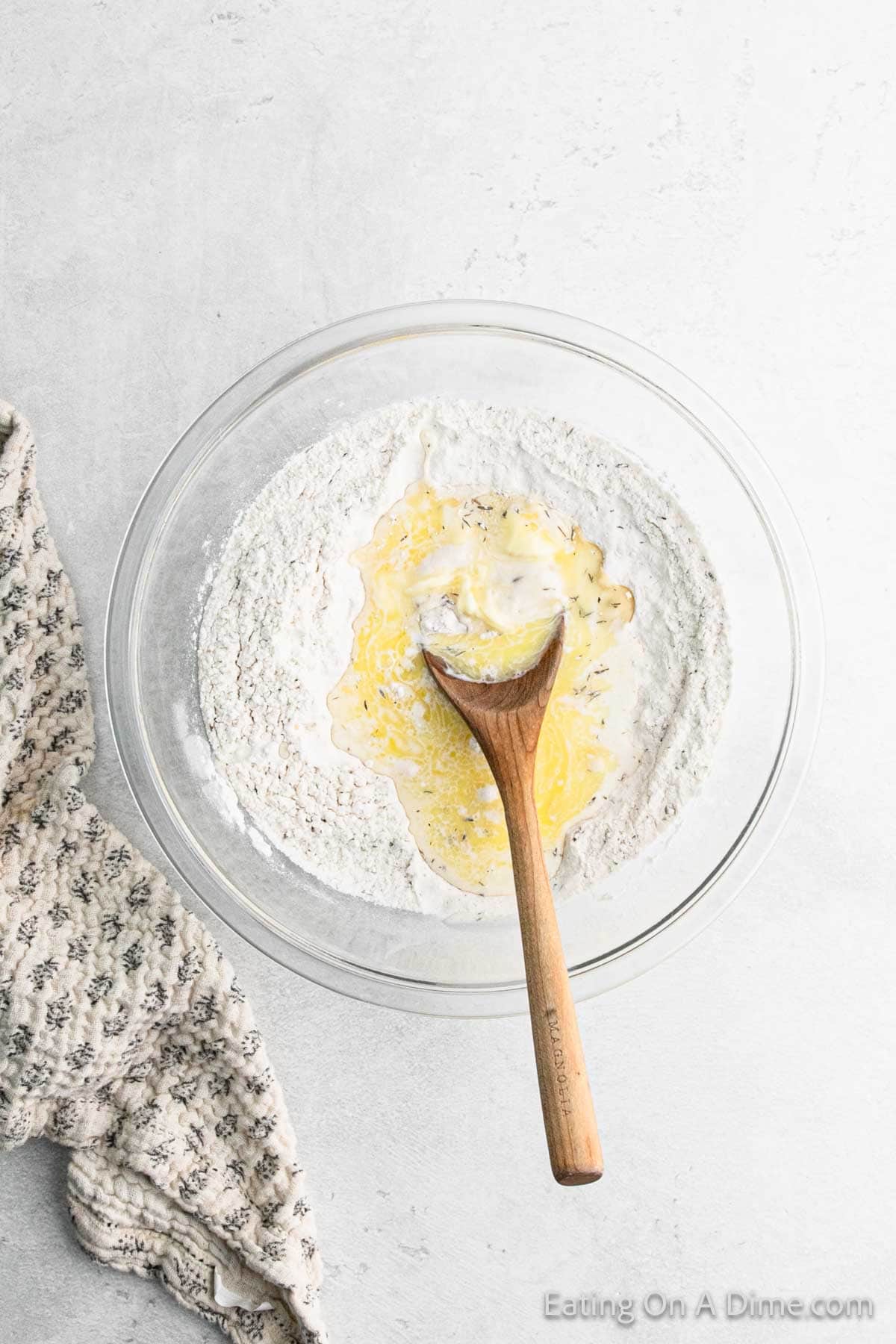 A glass bowl containing flour, melted butter, and herbs for a savory dumpling recipe, with a wooden spoon partially mixing the ingredients. A patterned cloth is nearby, all set on a light gray surface.