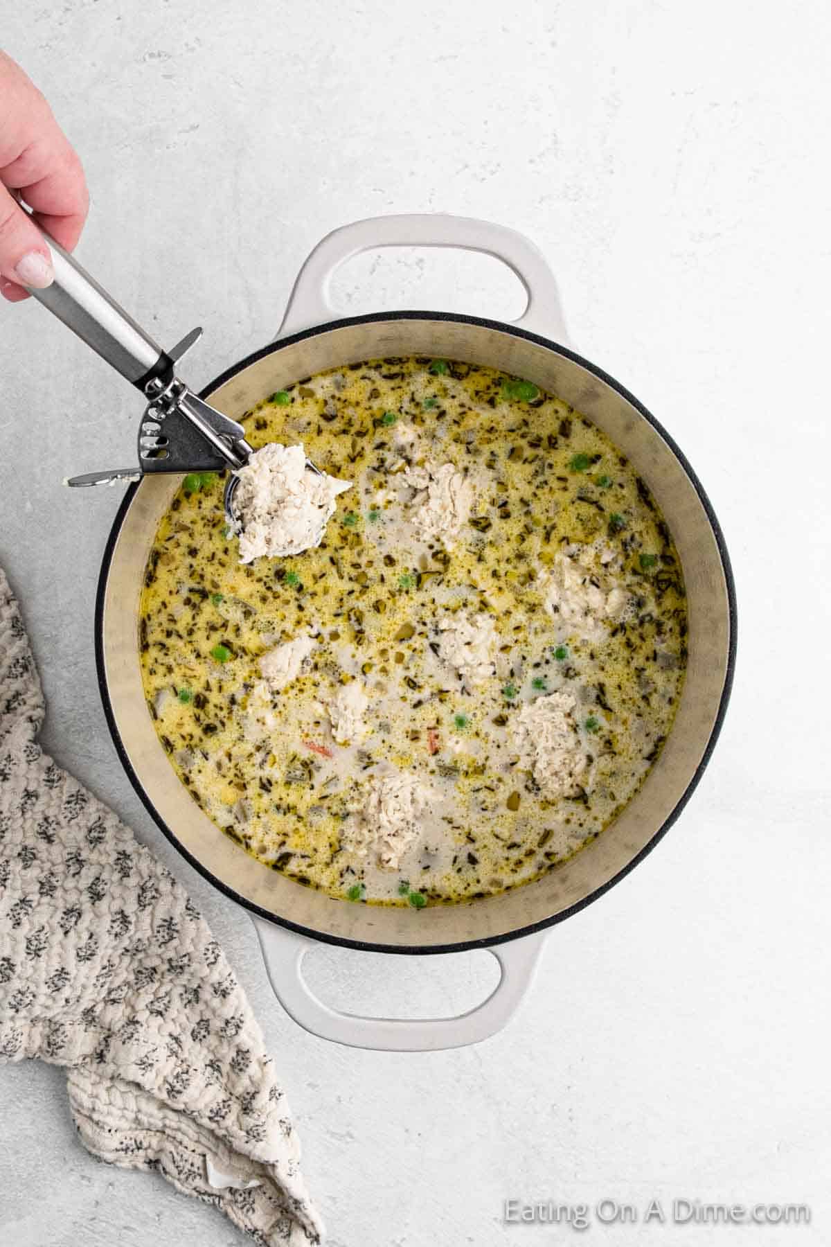 A large pot brims with creamy, speckled soup featuring chunks of chicken and herbs. A hand holds a scoop of raw meat above the pot, ready to be added. Dumplings bob gently in the broth, while a patterned cloth is partially visible on the side.