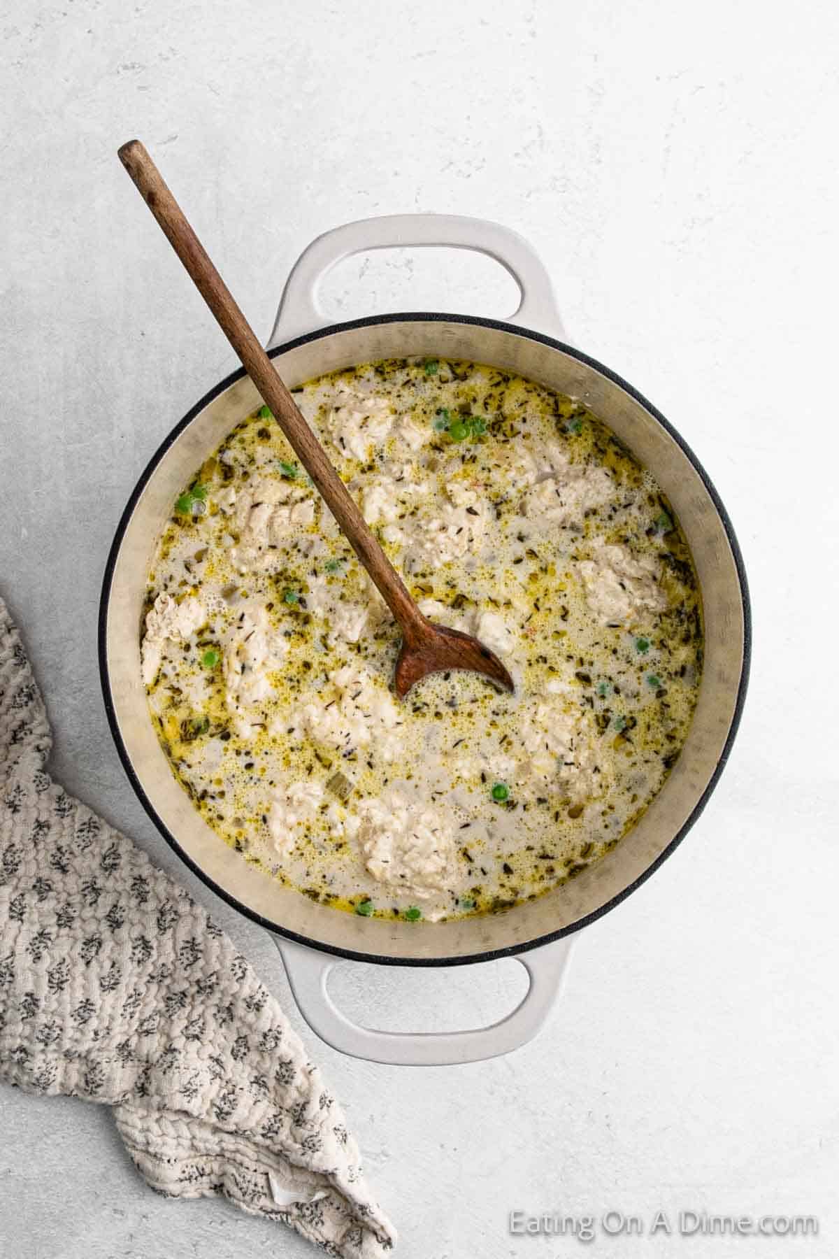 A large pot brims with creamy chicken and dumpling soup, mingled with herbs and small vegetables. A wooden spoon rests inside, while a textured cloth lies nearby on a light gray surface.