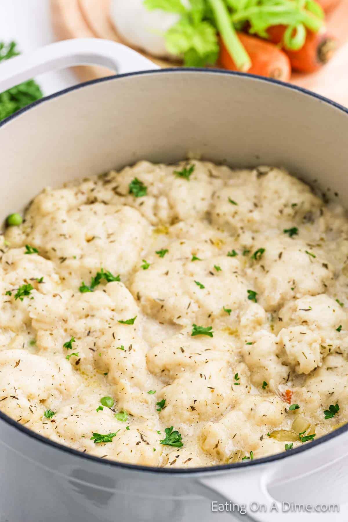 A white pot brimming with a comforting chicken and dumplings recipe, garnished with fresh herbs. In the background, vibrant carrots and crisp celery await on a wooden cutting board, ready to elevate your next culinary creation.